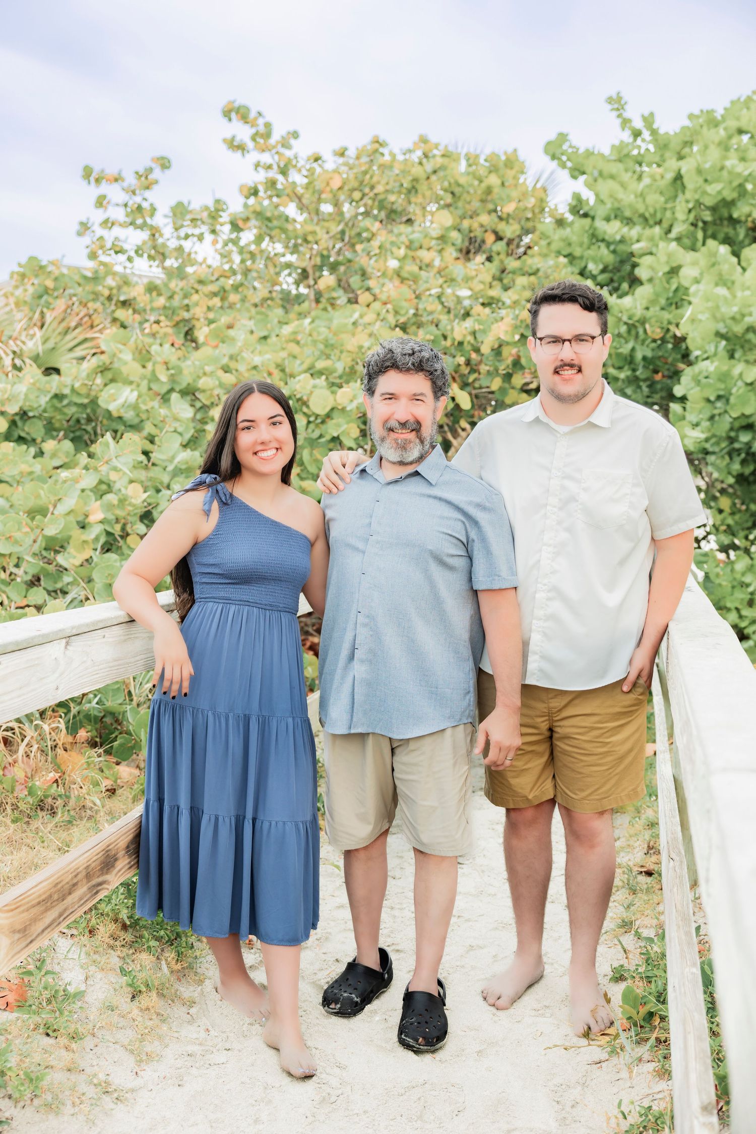 Three smiling people pose on a sandy beach boardwalk surrounded by lush green trees on a cloudy day.