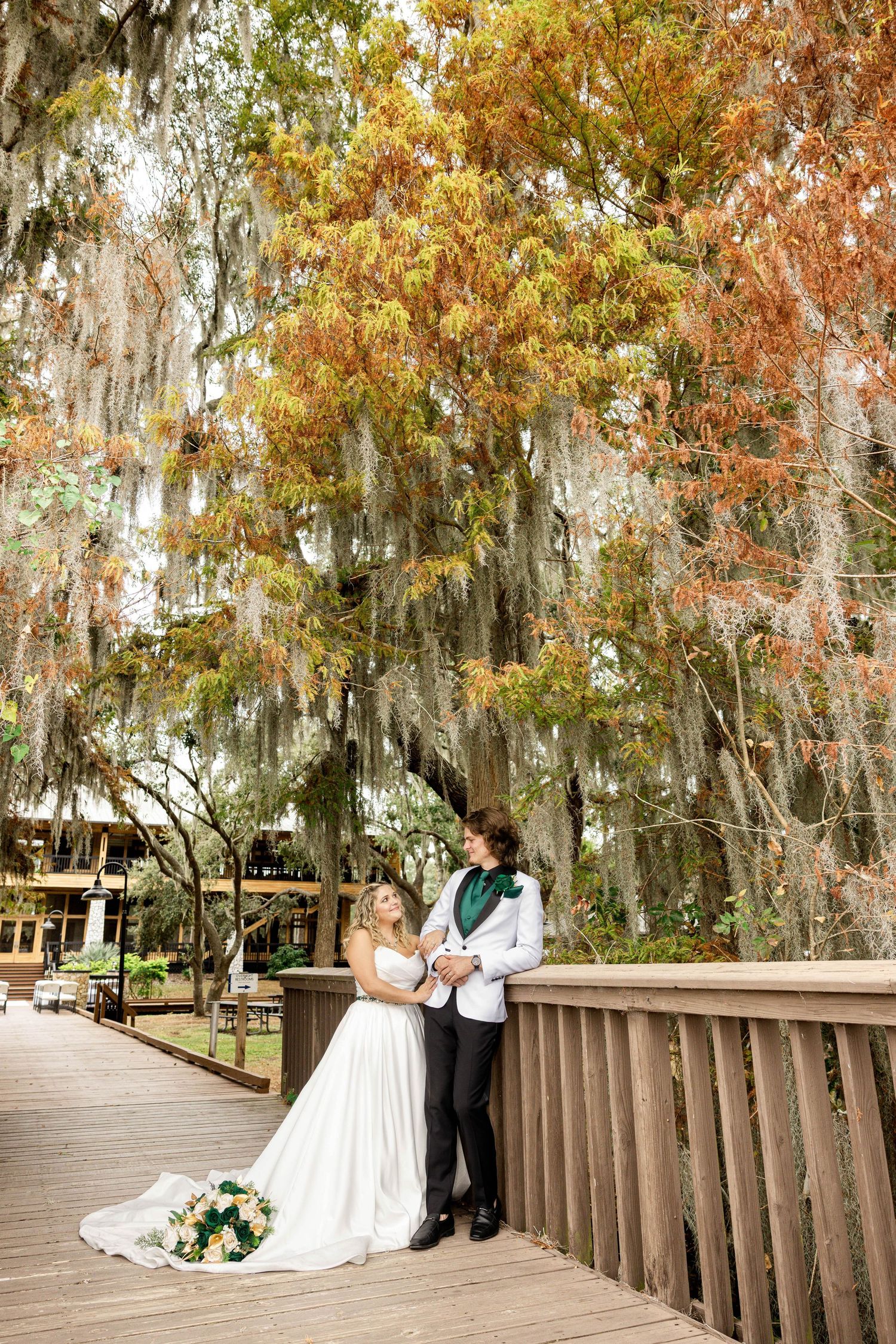 Bride and groom pose on wooden boardwalk under Spanish moss-draped autumn trees.