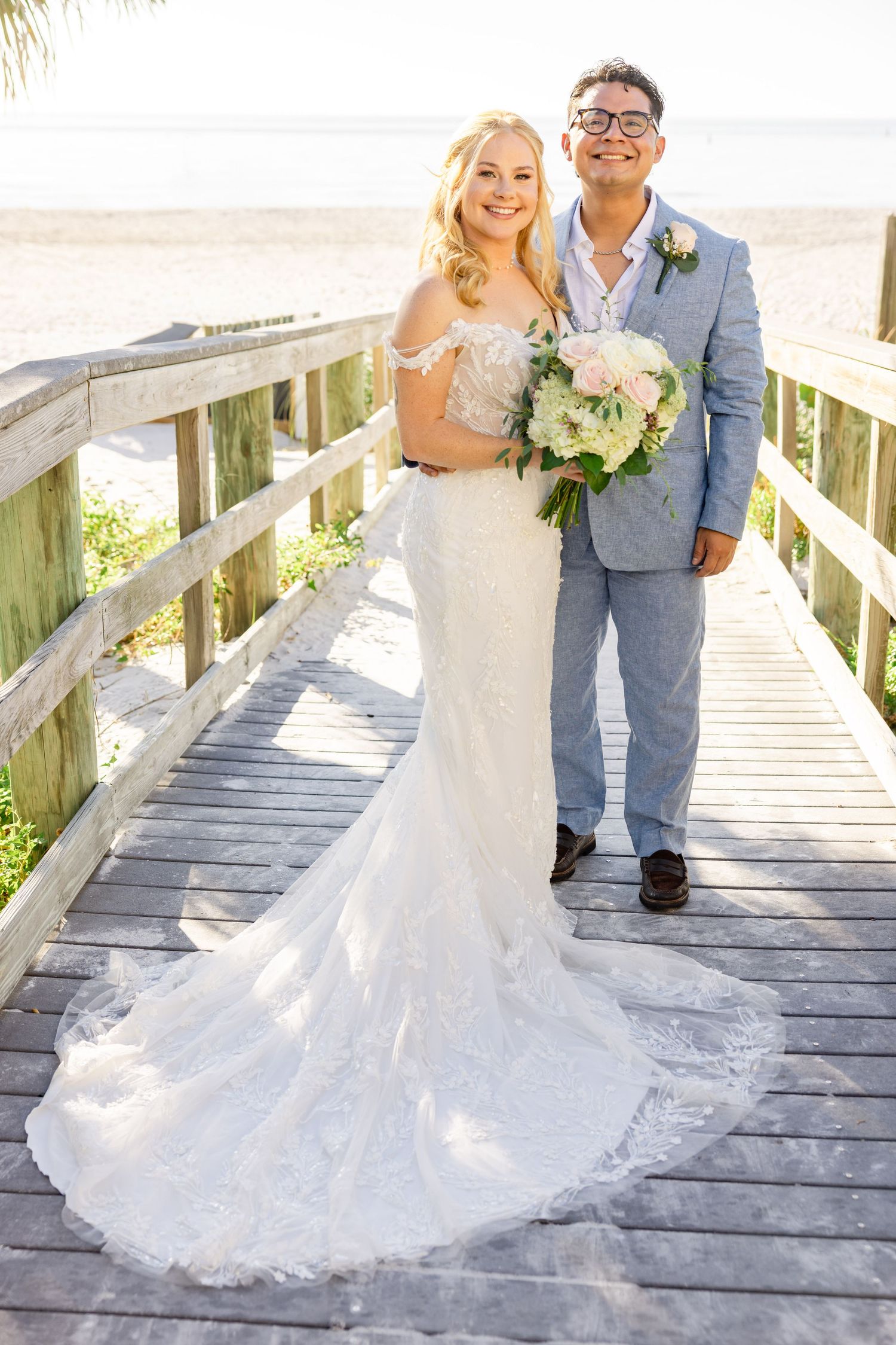 Bride in lace mermaid gown with long train and groom in blue suit pose on sunny beach boardwalk.