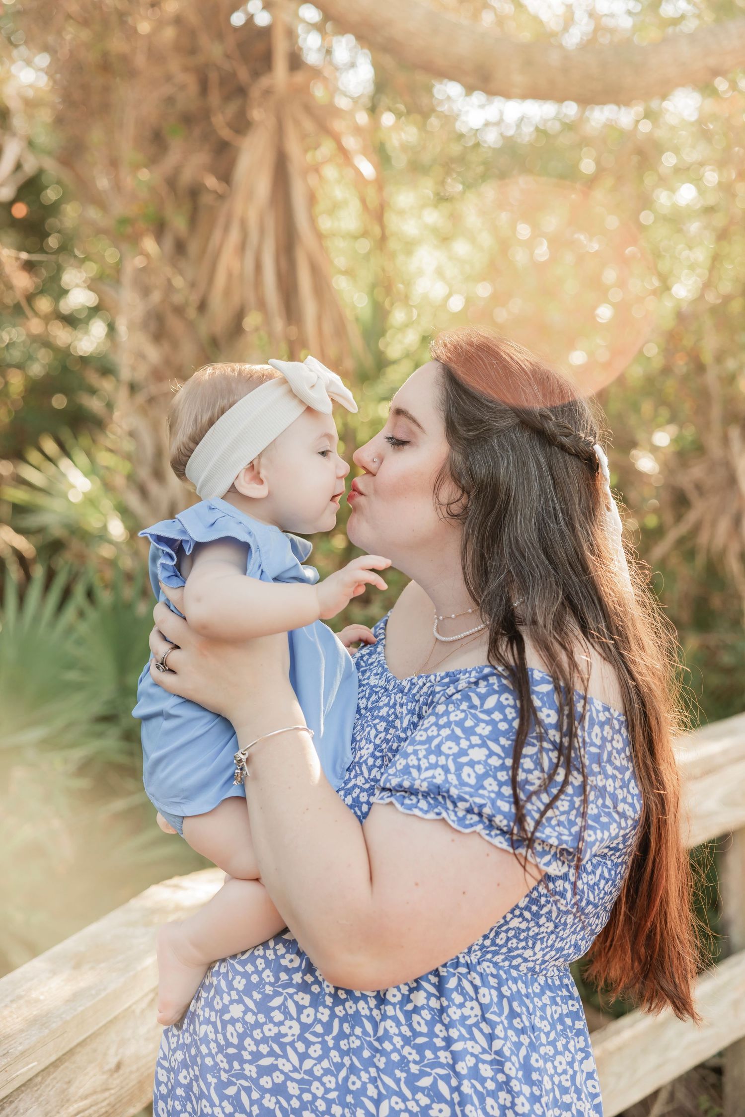 Mother kissing baby girl in blue dress outdoors in golden sunlight.