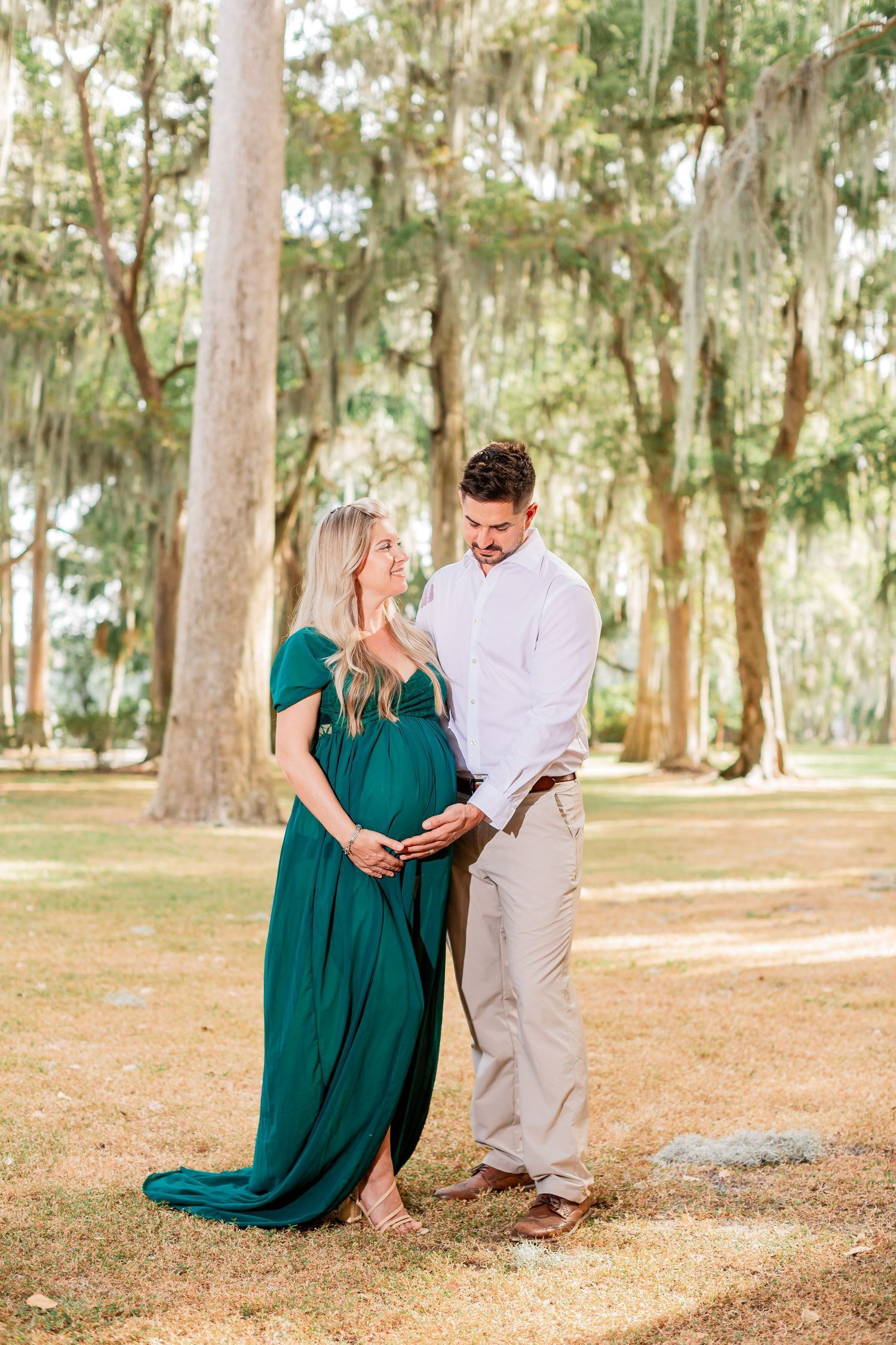 Couple in green and white outfits share tender maternity moment in park.