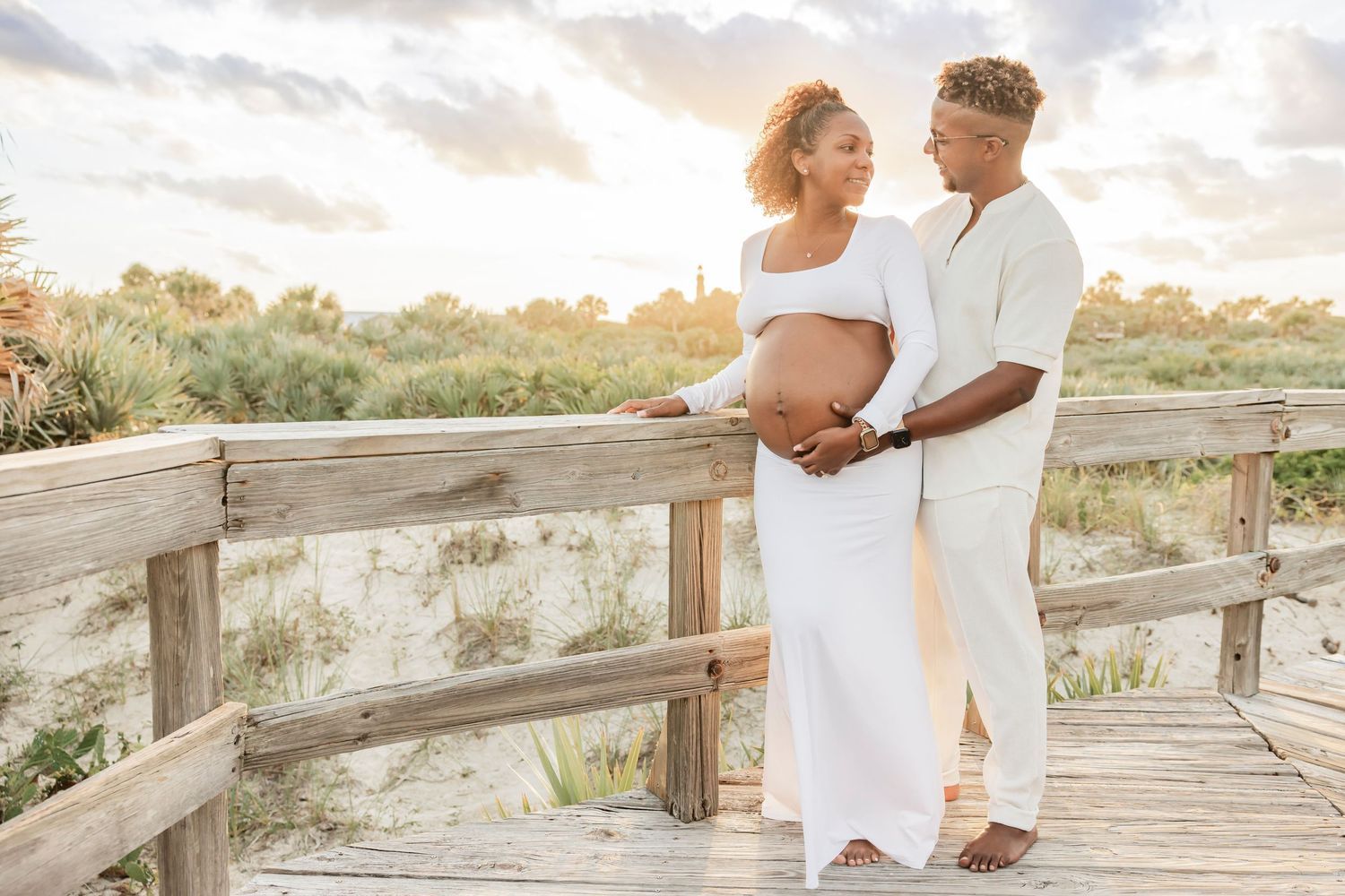 Expecting couple embracing on a wooden boardwalk surrounded by coastal dunes at golden hour.