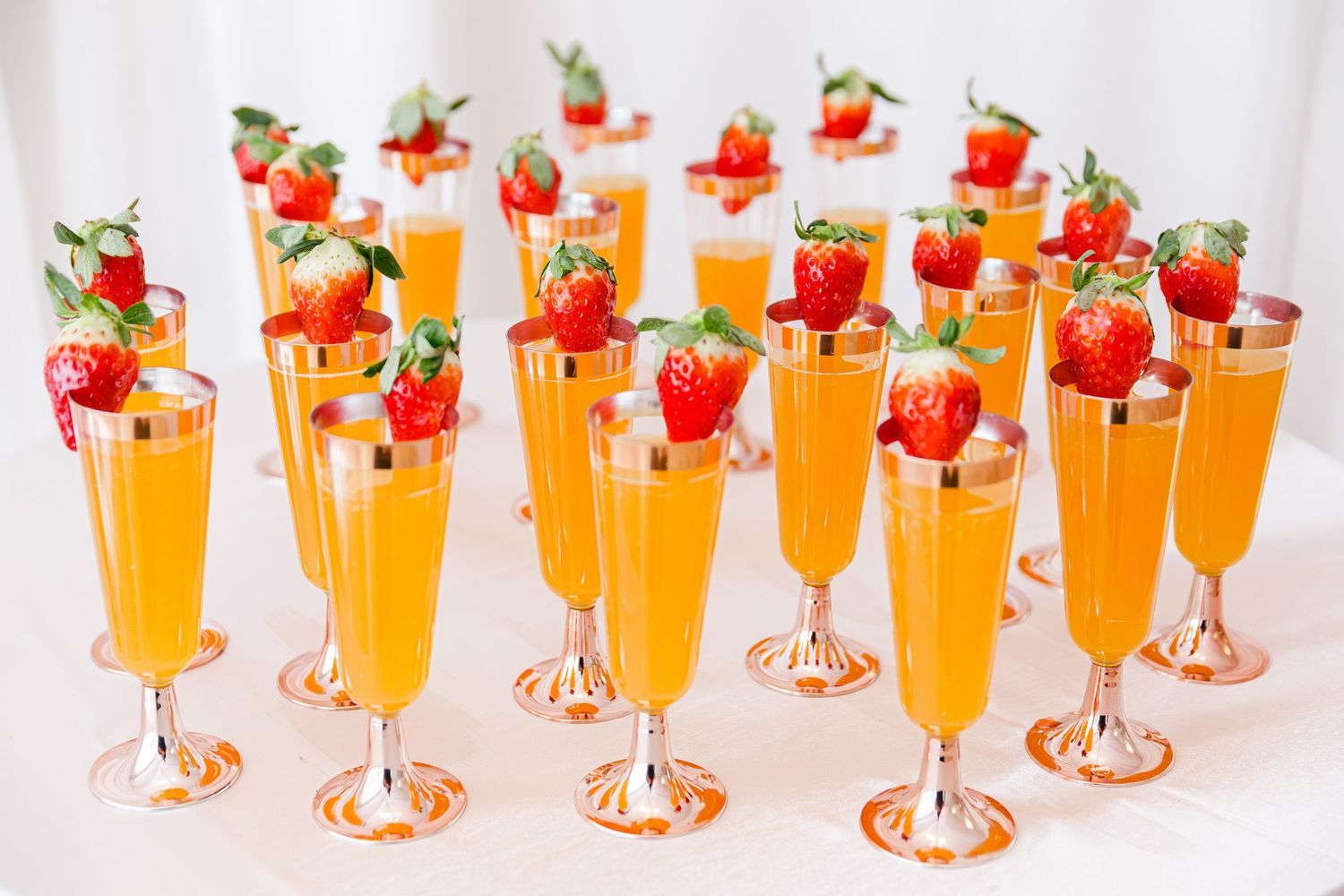 Elegant champagne flutes filled with orange juice topped with fresh strawberries on white table.