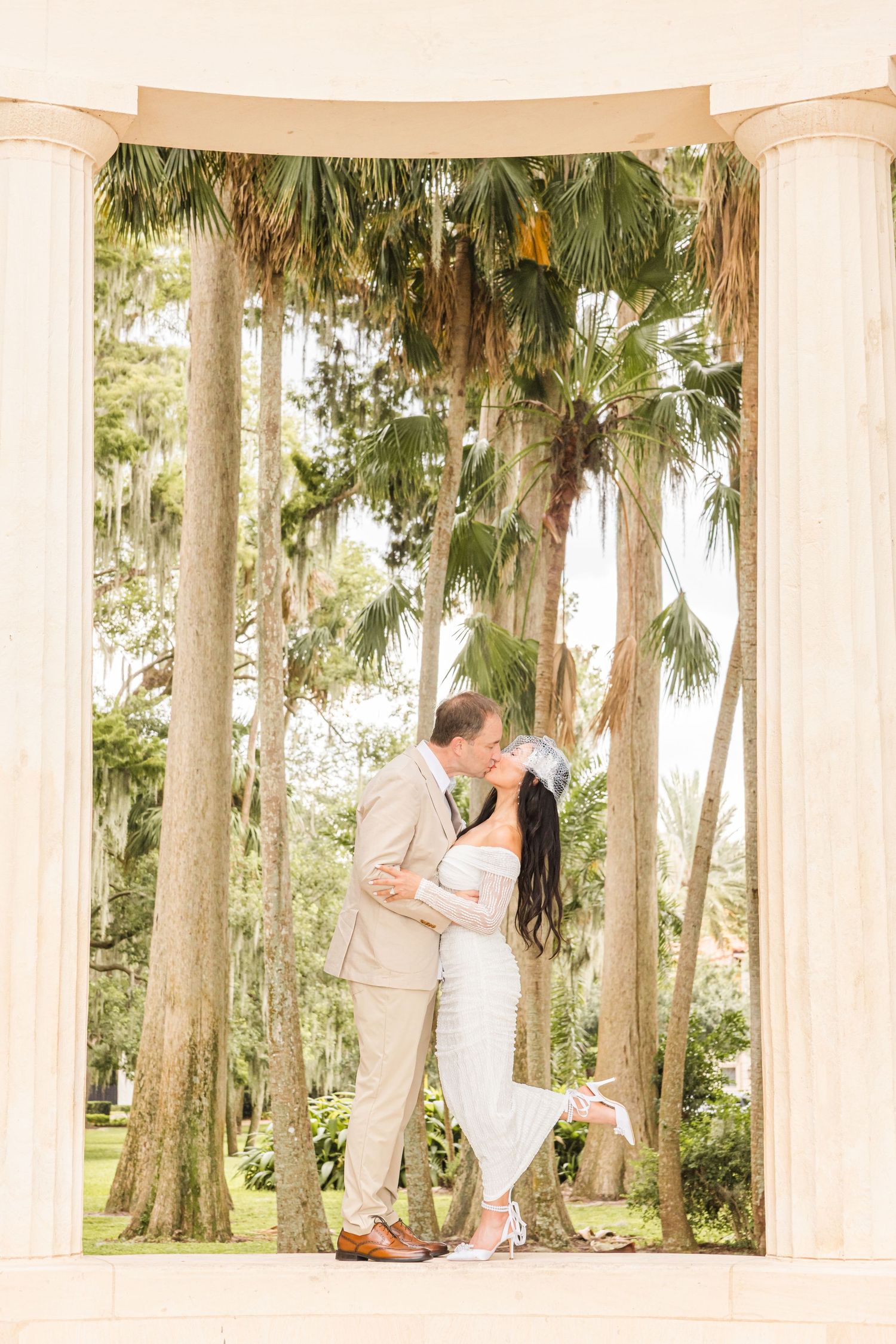 Couple shares romantic kiss between columns surrounded by palm trees at outdoor wedding.