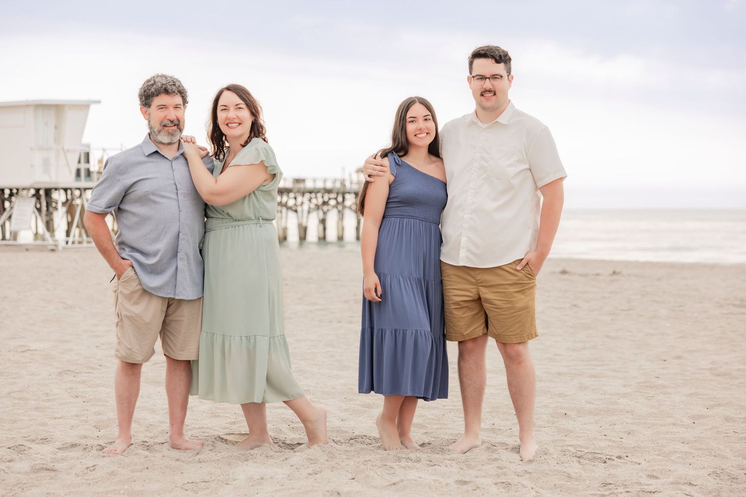 Family of four smiling together on a sandy beach near a pier on a cloudy day.