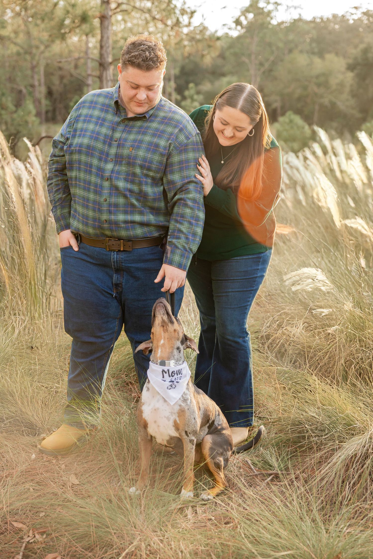 Couple petting their dog wearing a Mom bandana while standing in tall grass at golden hour.