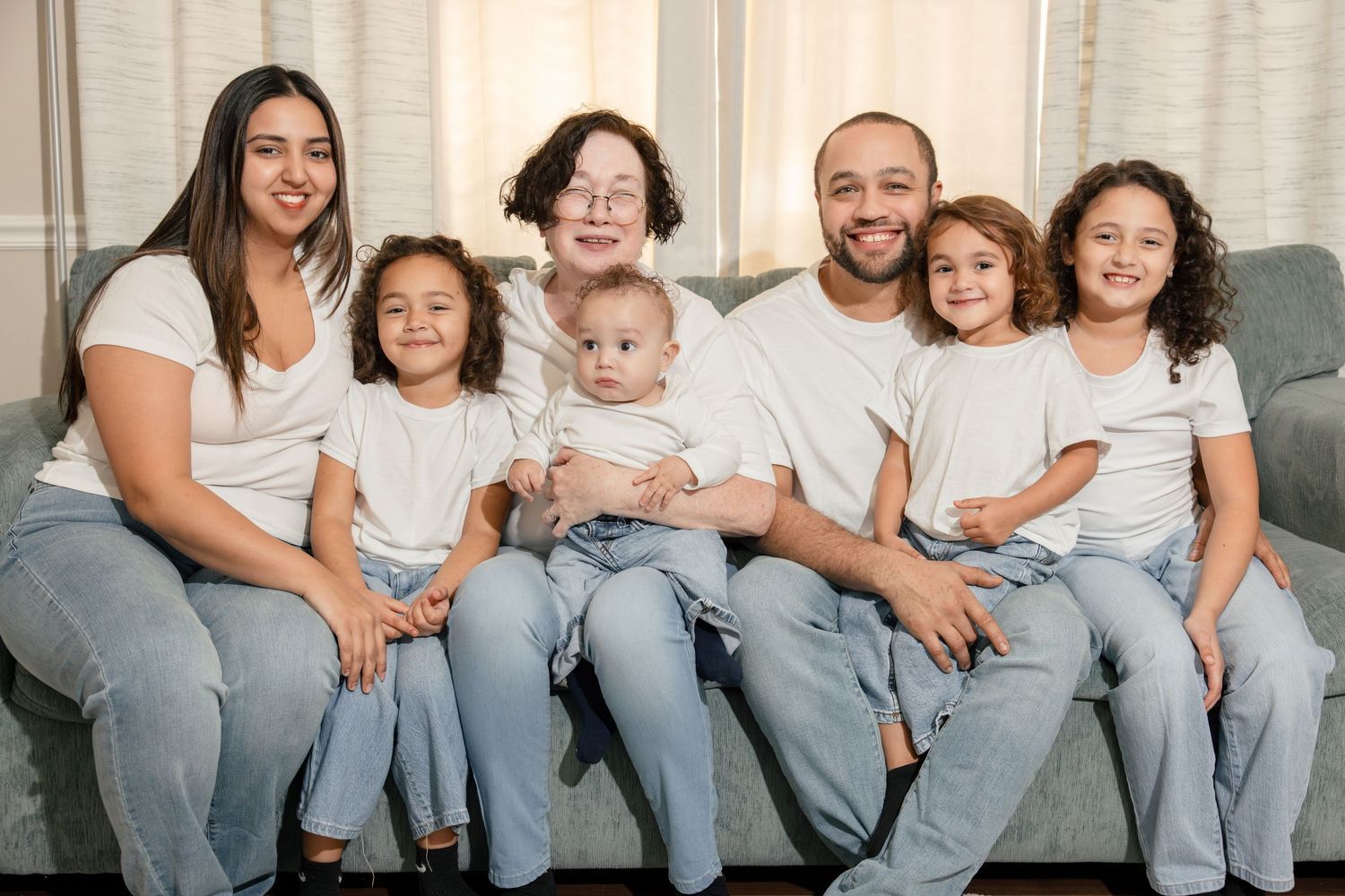 Extended family of seven in white tops and jeans posing together on a couch in a bright living room.