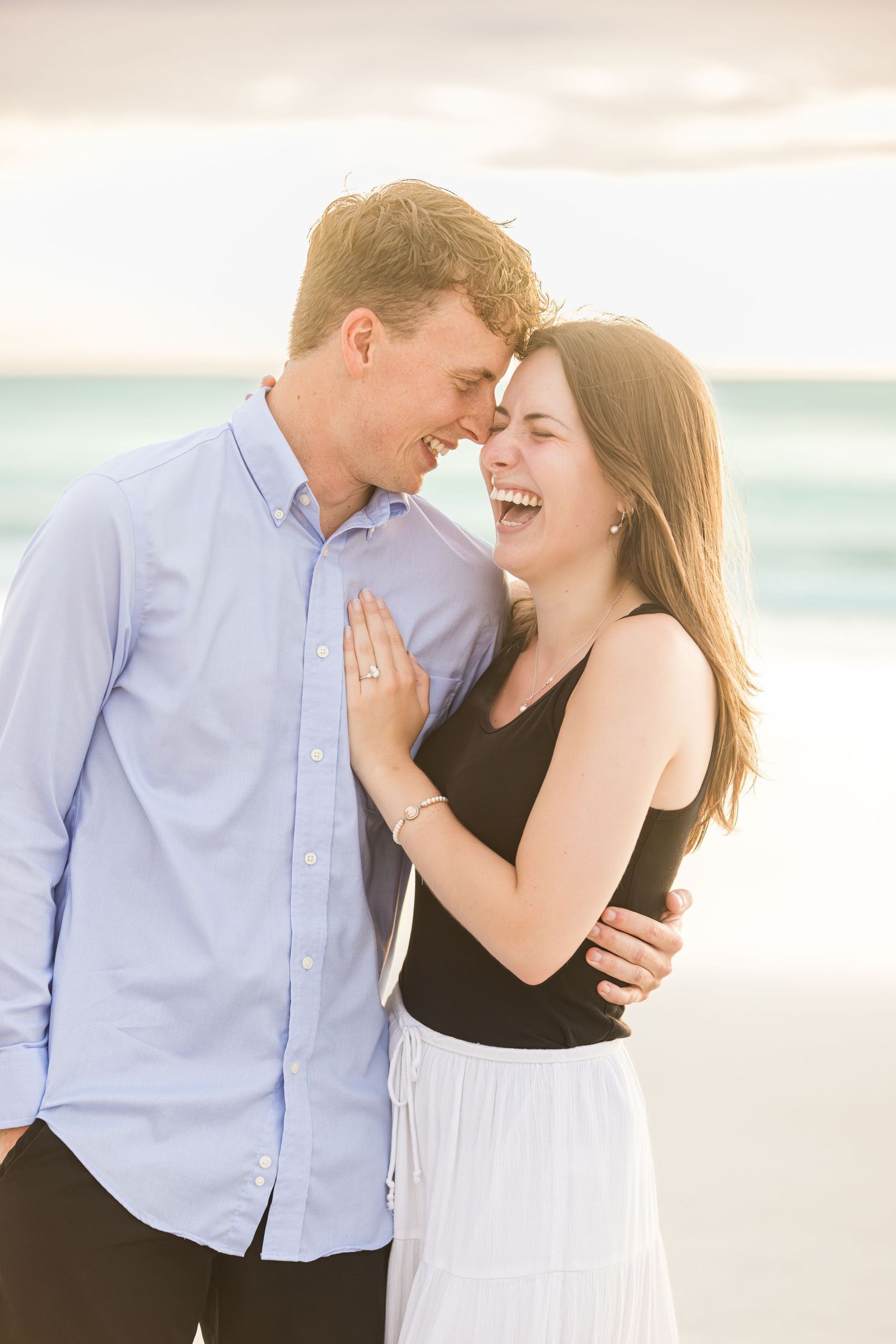 Happy couple laughing together on a beach at sunset, woman showing engagement ring in romantic photo.