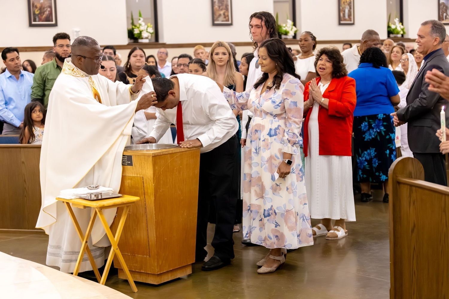 A priest performs a baptism ceremony at the altar of a church filled with congregation members in white attire.