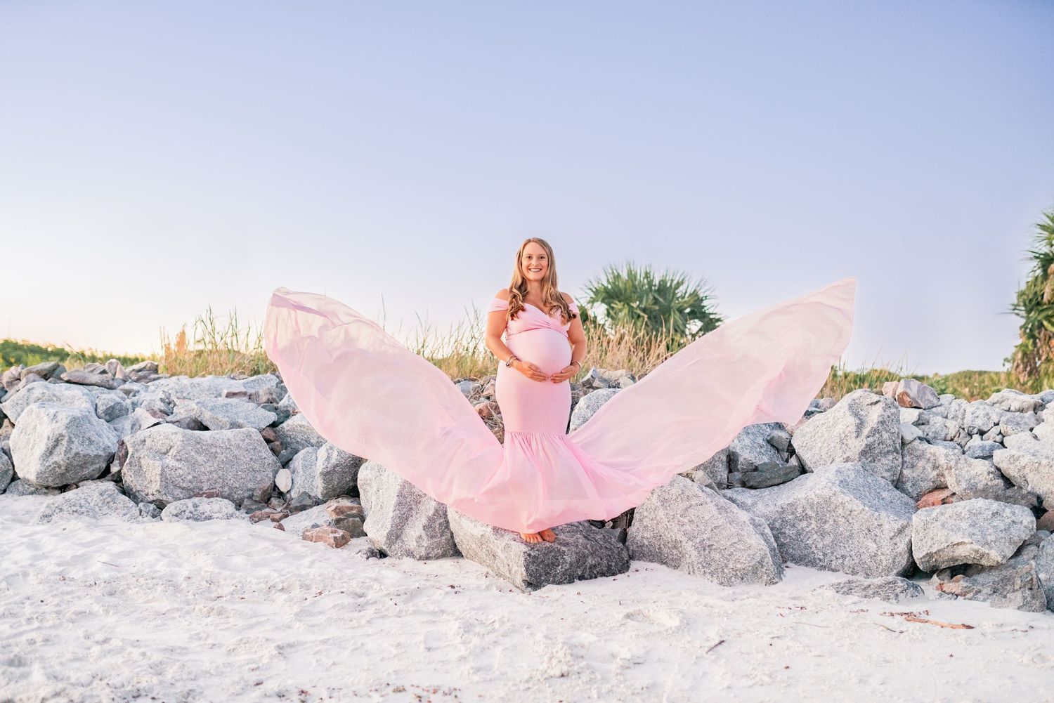 Pregnant woman in pink flowing dress twirls on rocky beach at sunset.