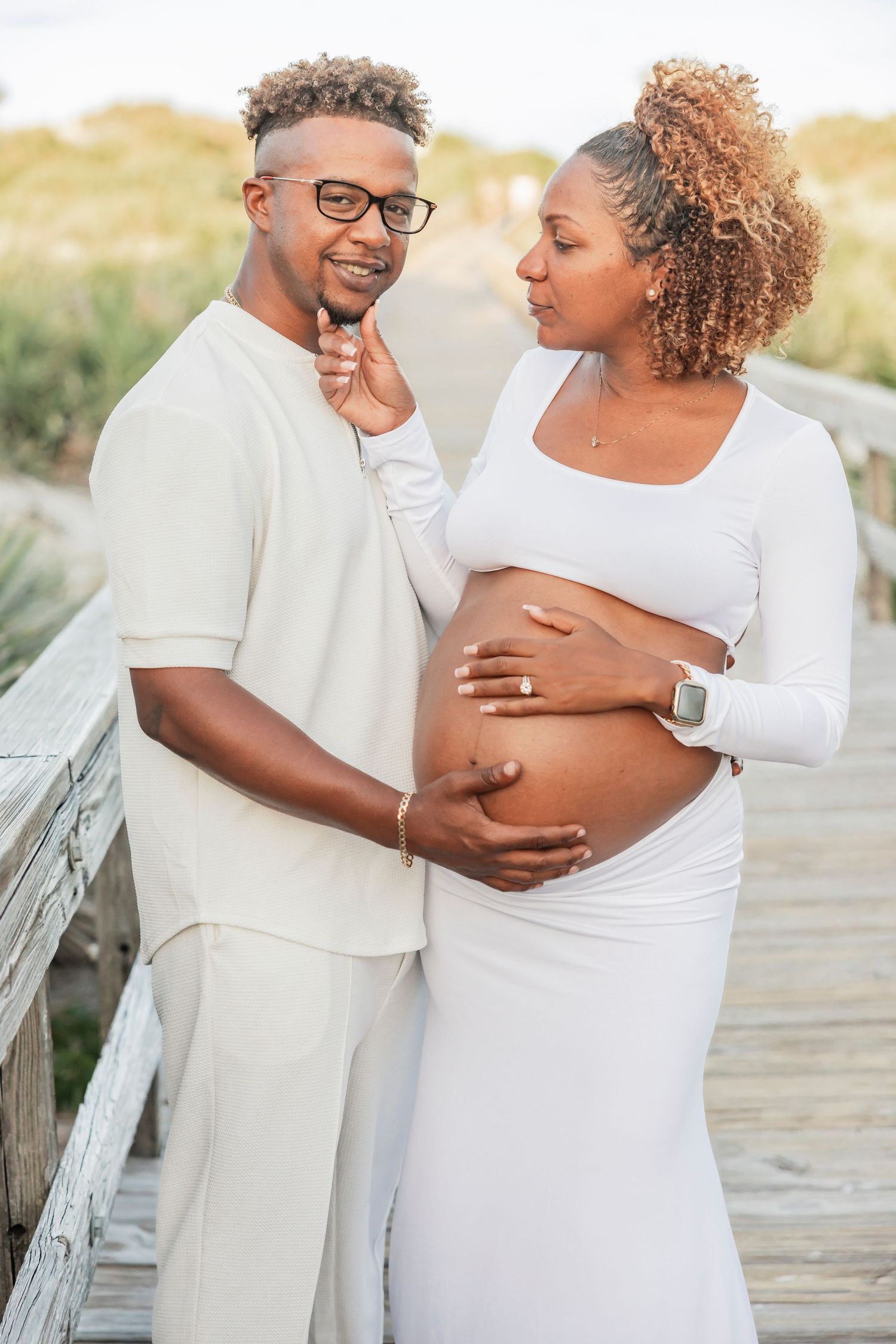 Couple on a sandy boardwalk, man smiling at camera while pregnant woman in white dress holds her baby bump.