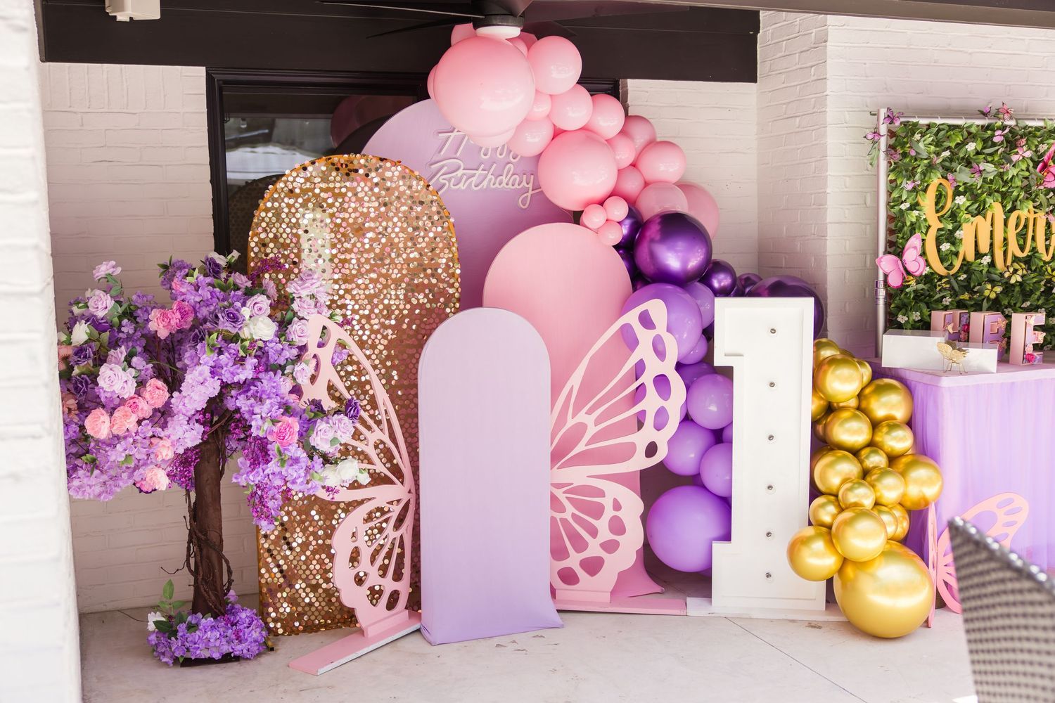 Pink purple and gold butterfly themed birthday party decoration setup with balloon arch sequin column and floral tree.