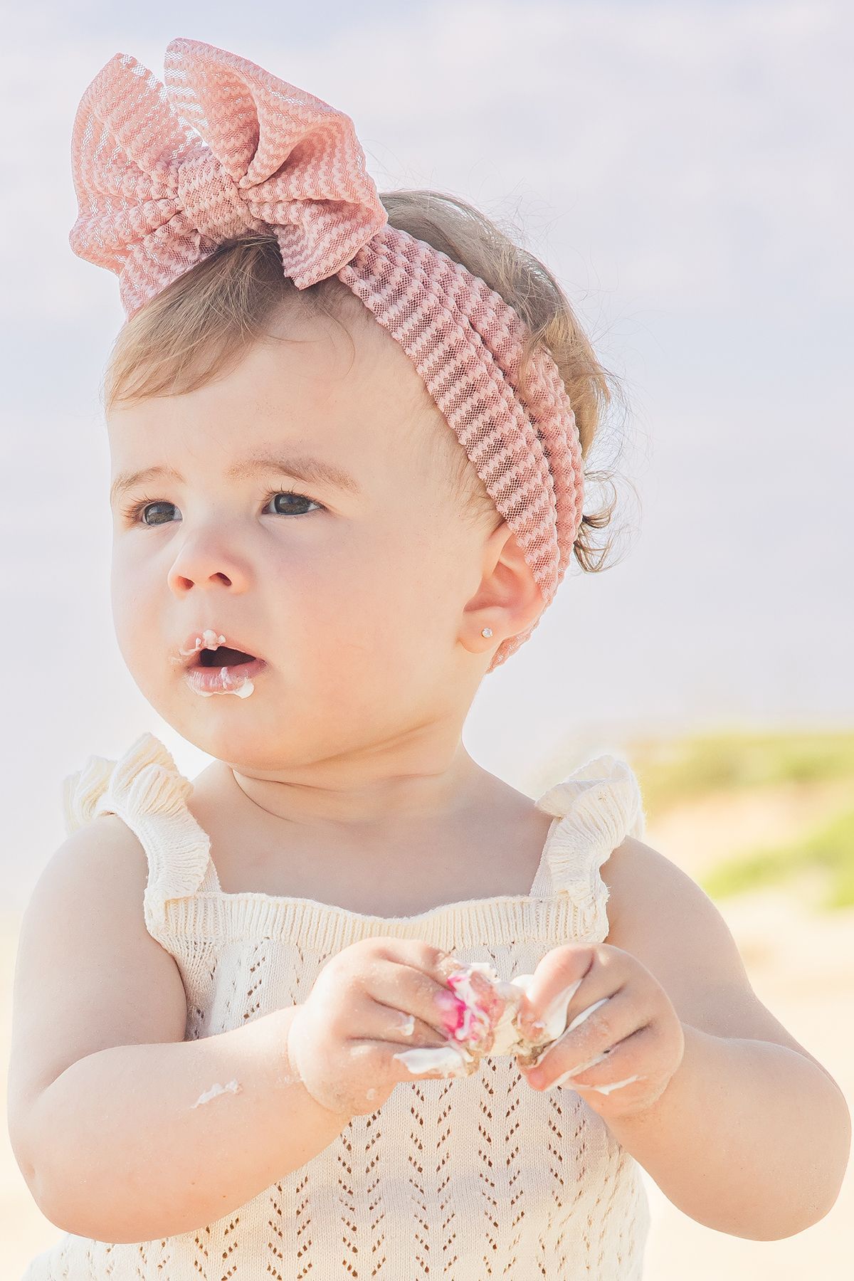 Cute baby girl wearing pink bow headband and white dress holding a small flower outdoors.