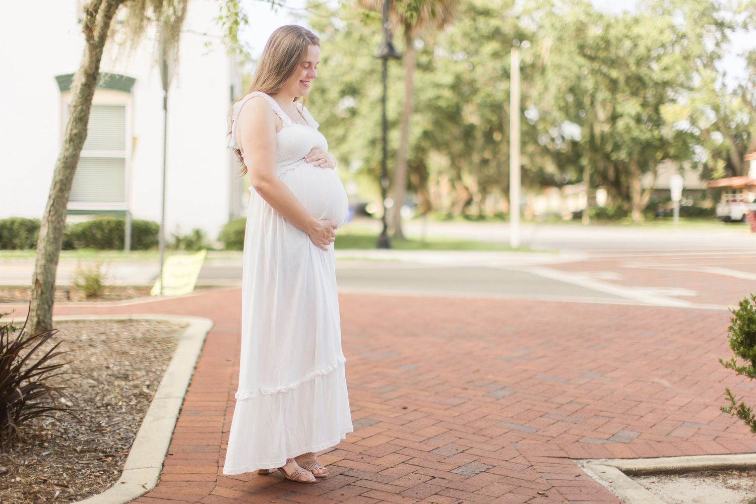 Pregnant woman in white maxi dress standing on brick pathway in sunny outdoor park setting.