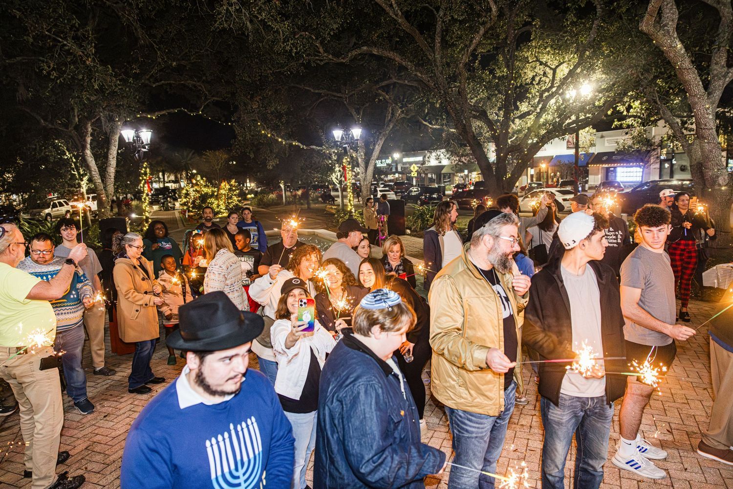 Community members holding sparklers and candles at outdoor Hanukkah celebration at night.