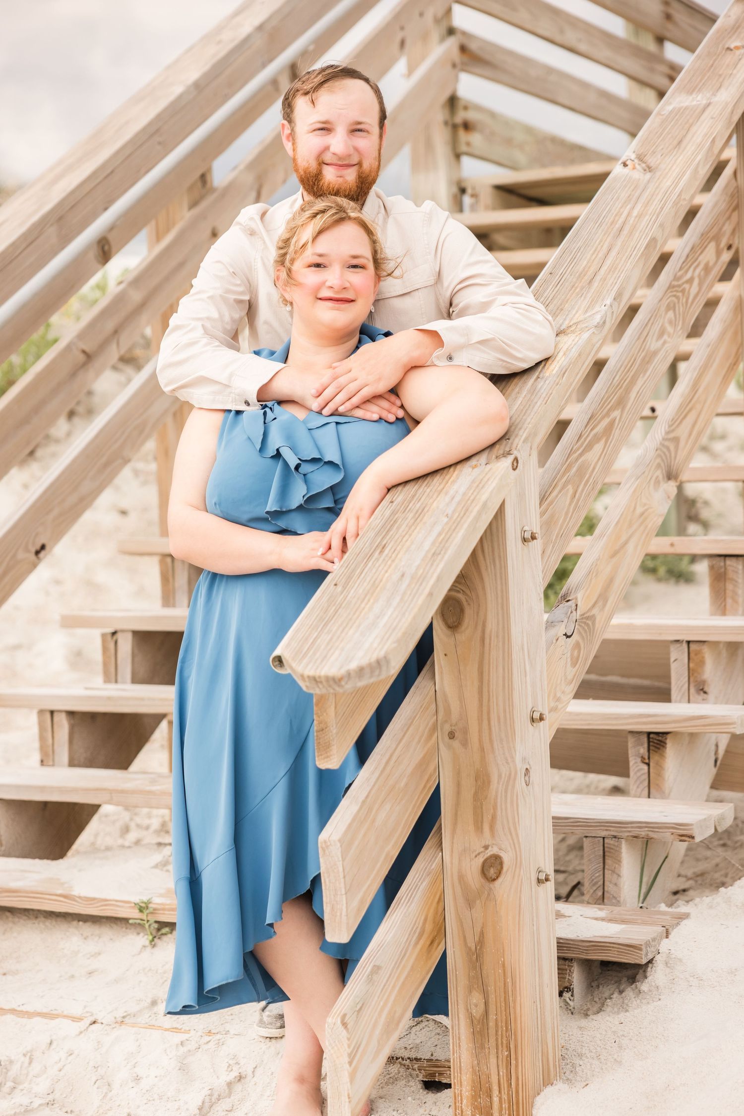 A couple poses on wooden beach stairs, man embracing woman in a blue dress from behind.