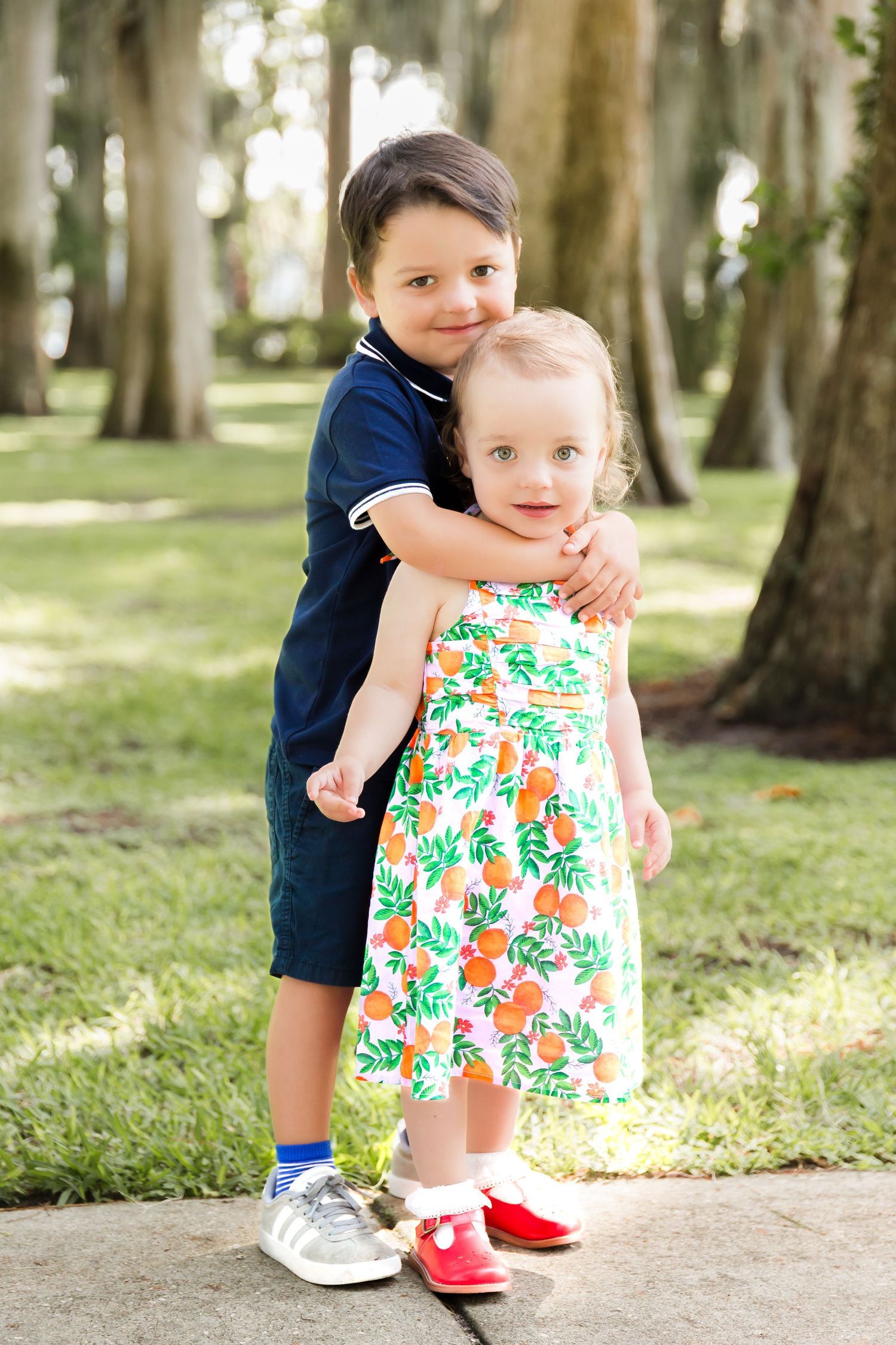 Young boy hugging his little sister in a floral dress while standing in a sunny park.