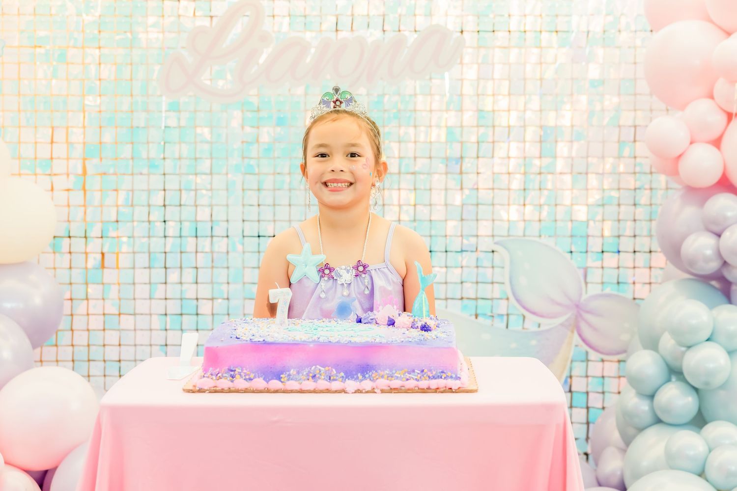 Smiling birthday girl wearing tiara standing behind mermaid cake at pastel balloon decorated party.