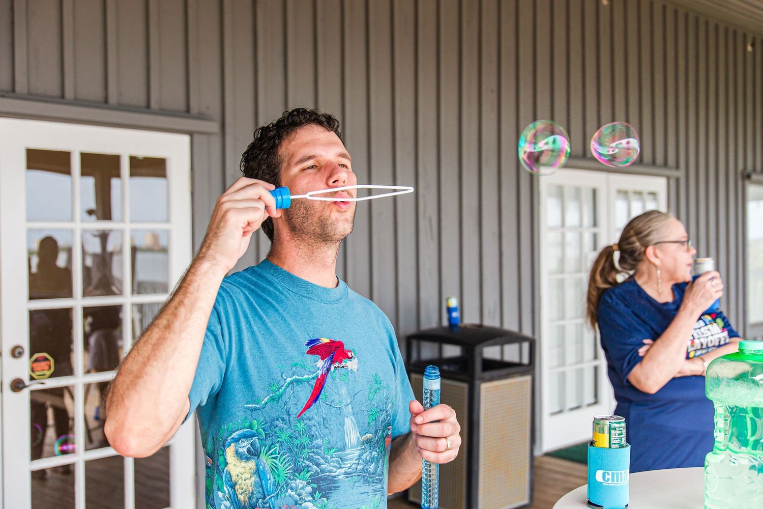 Man blowing bubbles outdoors on a covered porch at a casual daytime gathering.