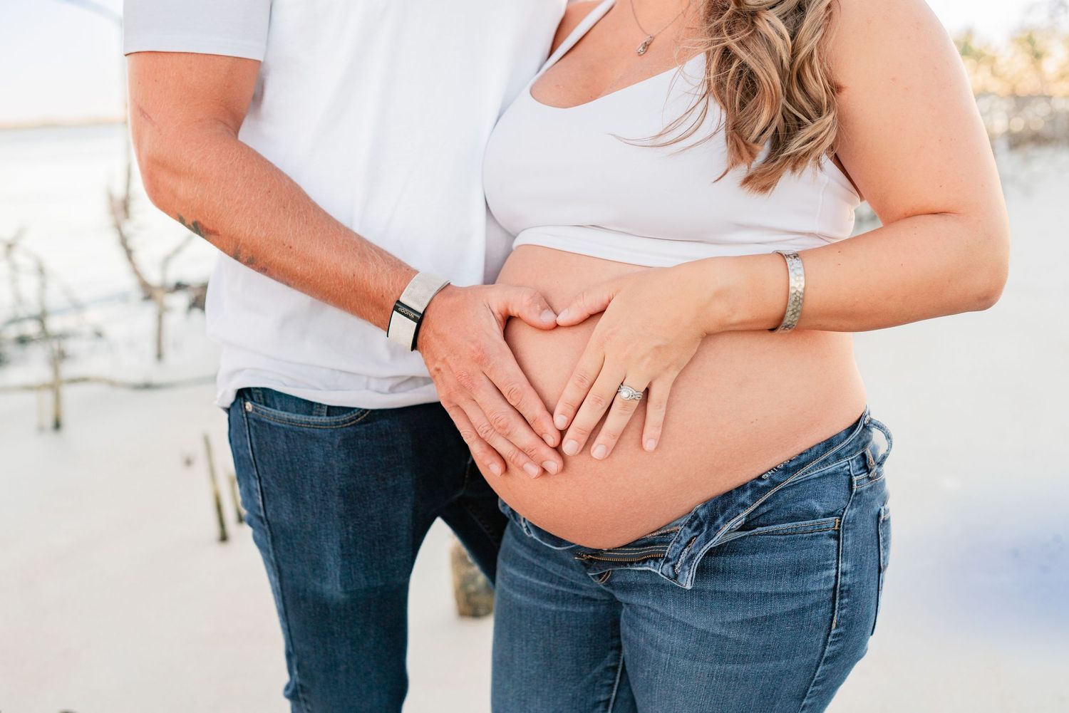 Couple forming heart shape with hands on pregnant belly, casual beach maternity photo.