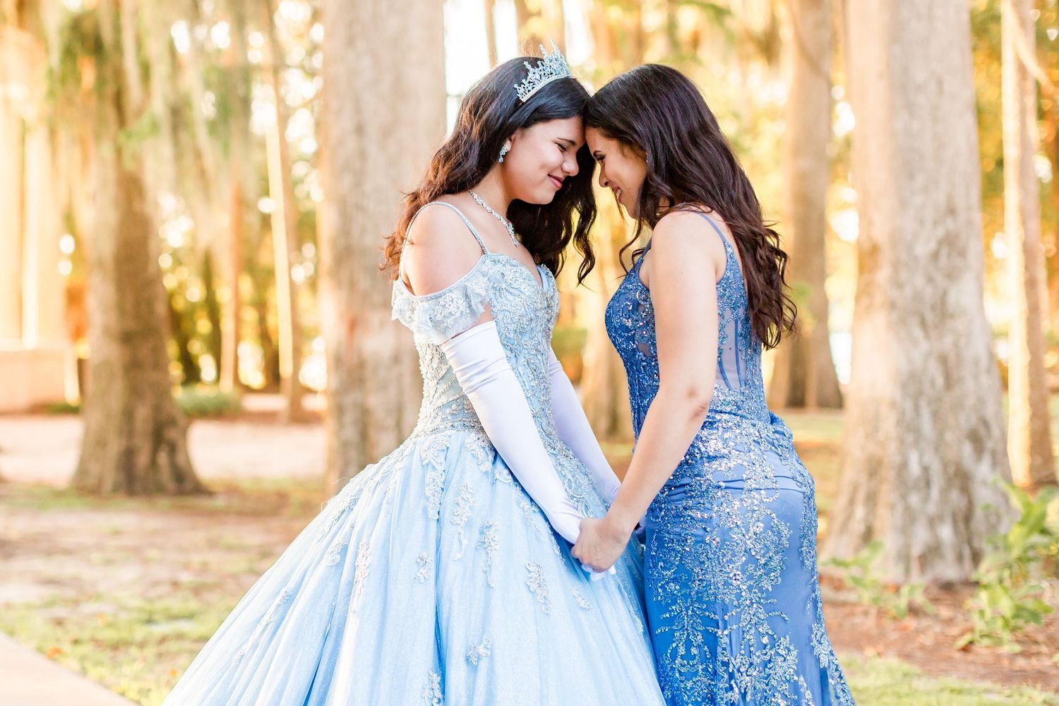 Two young women in matching blue formal gowns hold hands and face each other outdoors among trees.