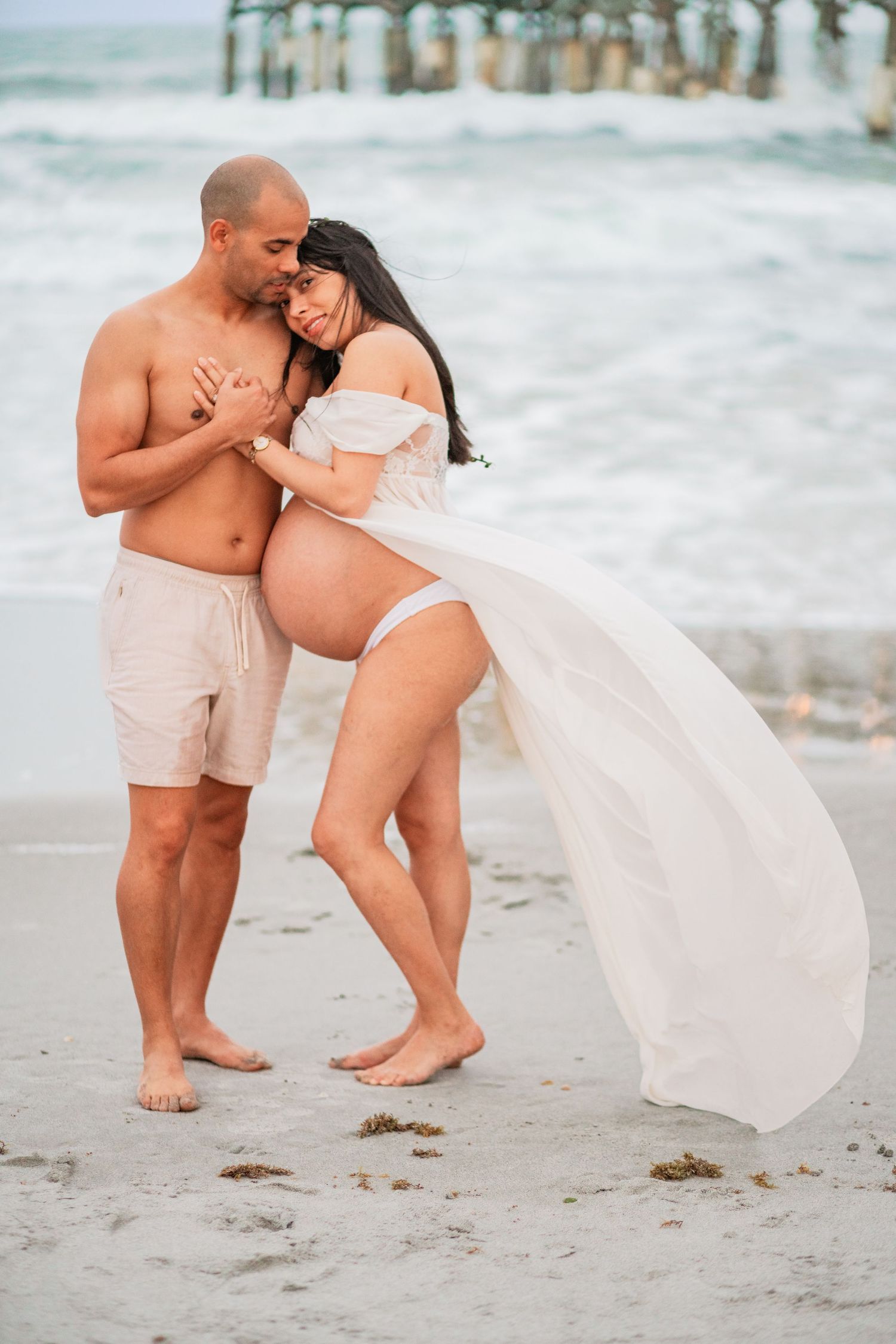 Couple embracing on beach at sunset, pregnant woman in flowing white dress with ocean backdrop.