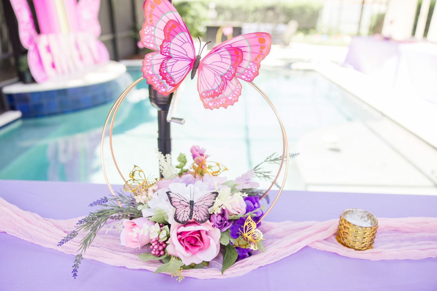 Elegant gold hoop centerpiece with pink butterfly and floral arrangement on a lavender table near a pool.