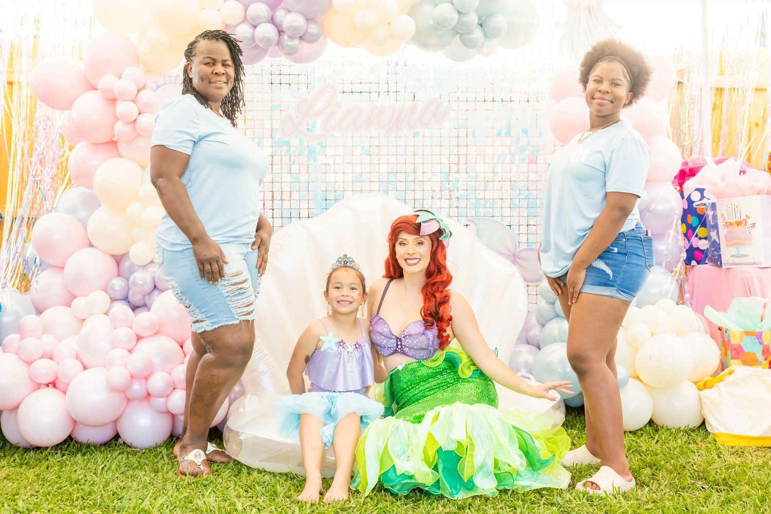 Two women and young girl posing with Ariel mermaid character at pastel balloon decorated party.
