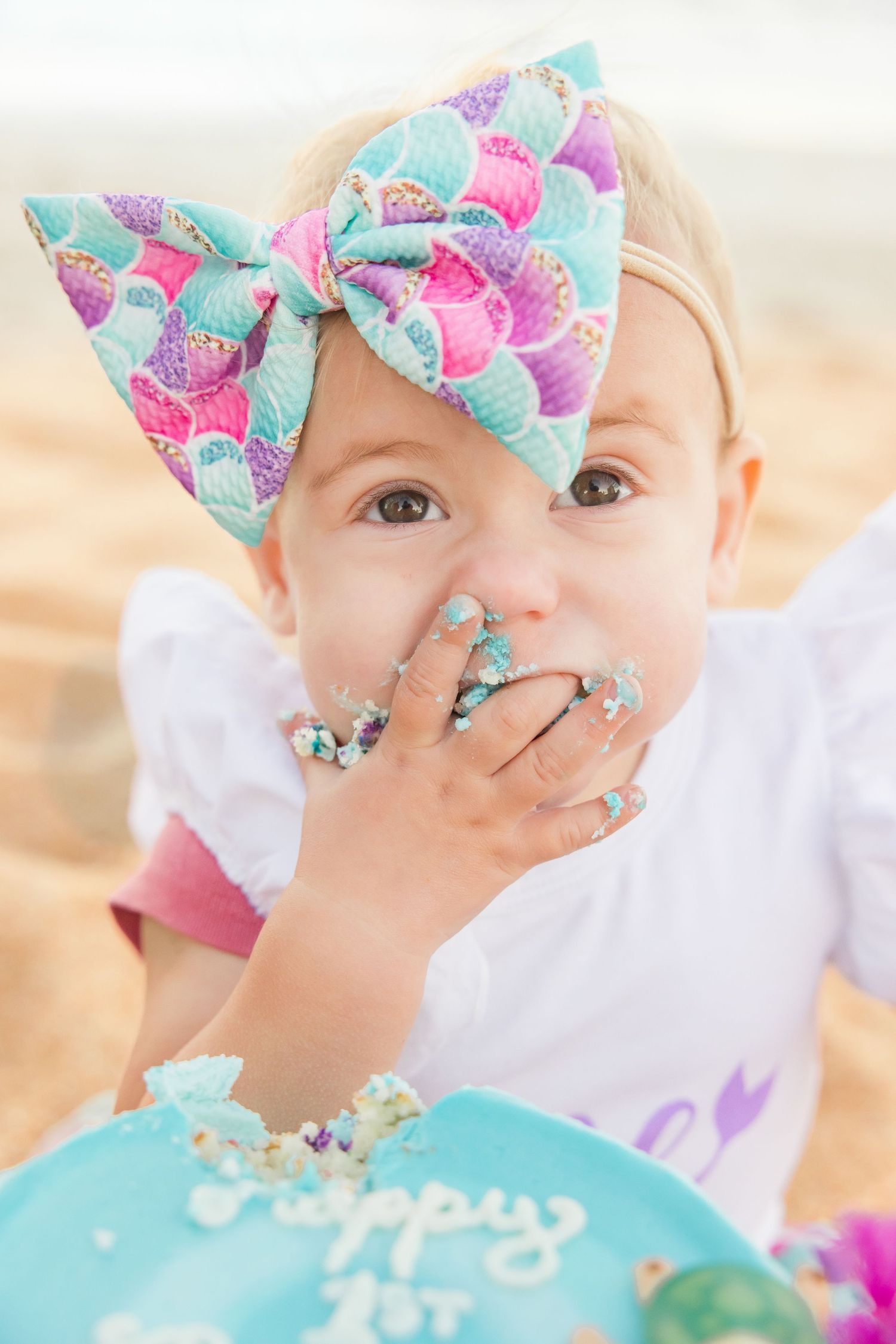 Close-up of baby girl with colorful mermaid bow eating birthday cake.