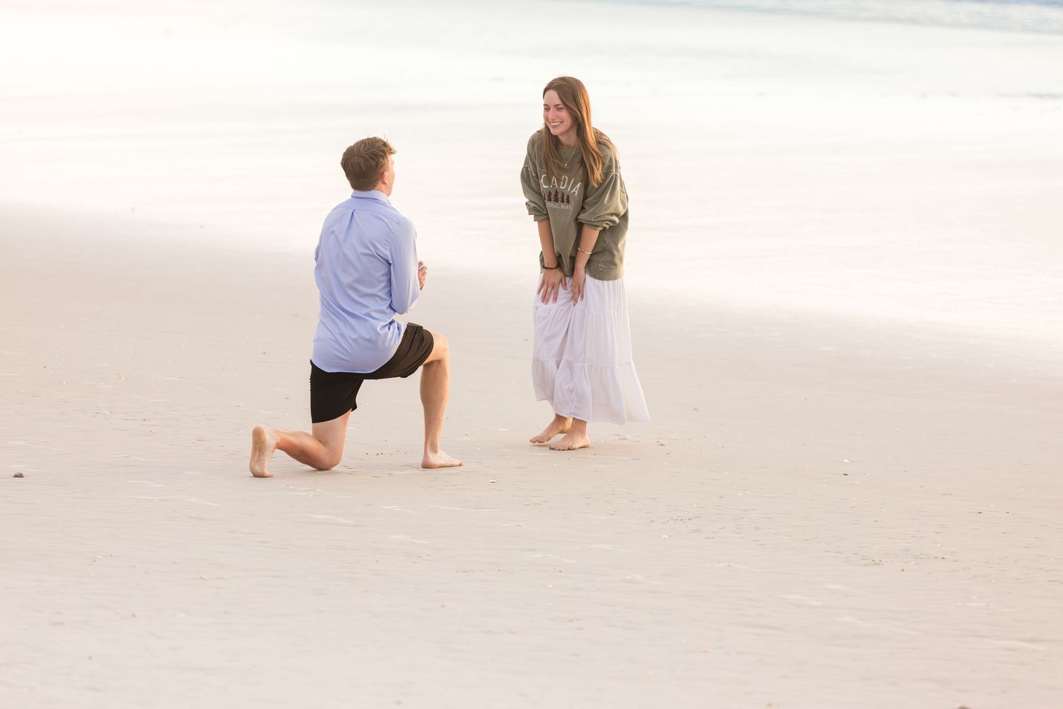 Man on one knee proposing to a woman on a white sandy beach at sunset.