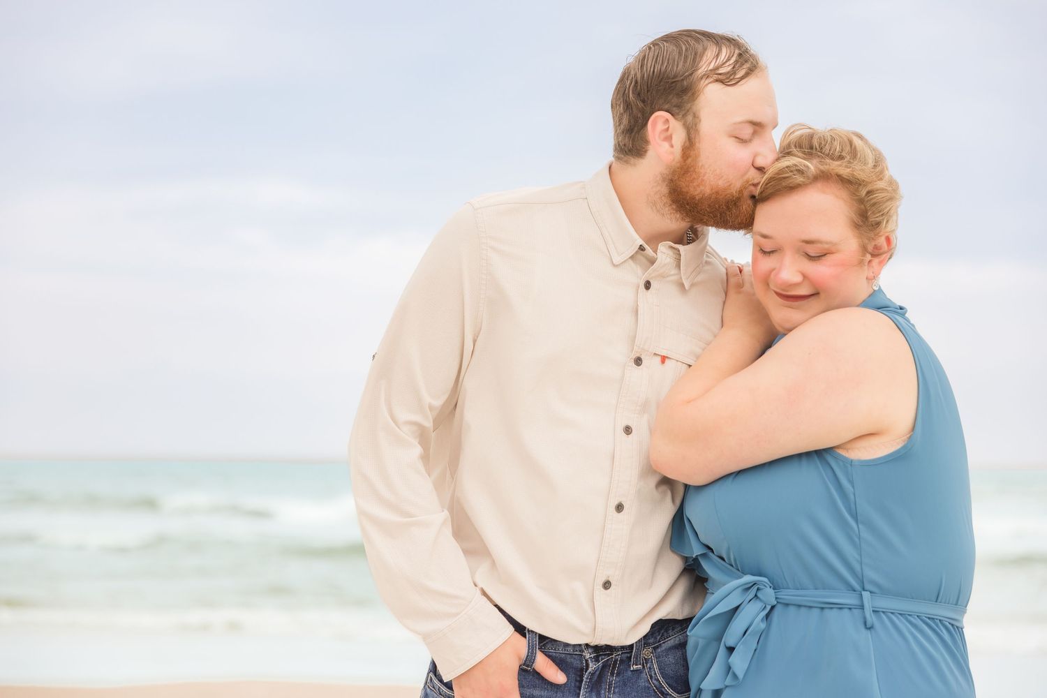 A bearded man kisses a blonde woman on the cheek as they embrace on a beach.