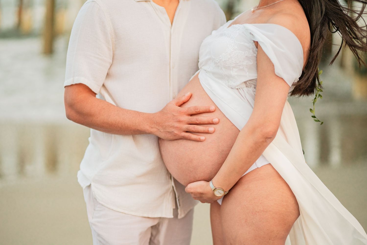 Couple in white outfits tenderly holding pregnant belly on beach, maternity photo session.
