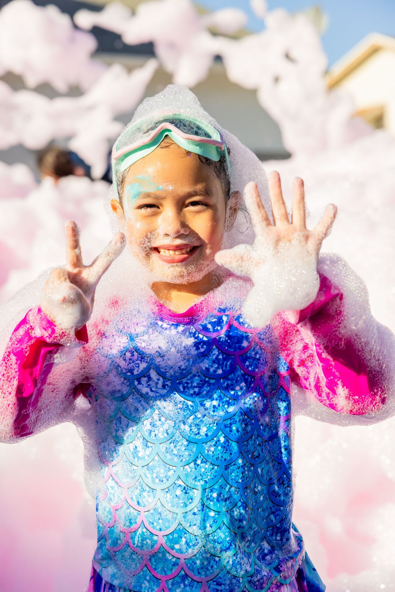 Excited young girl in mermaid costume covered in foam bubbles at outdoor foam party.