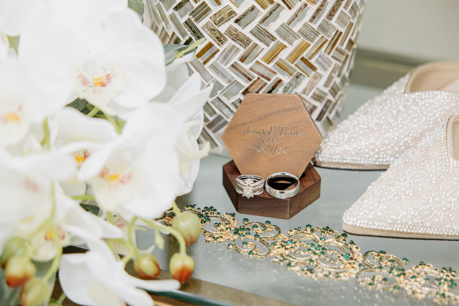 Wedding rings in a wooden box beside white orchids, a glittery heel, and gold jewelry on a mirrored tray.