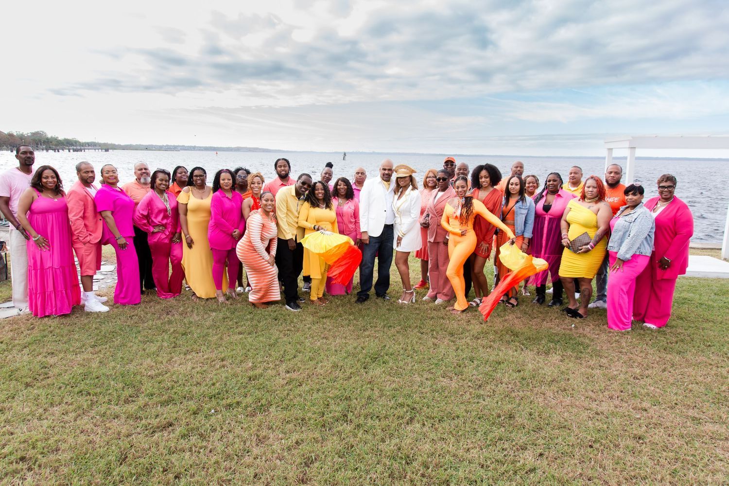 A large group of guests in colorful pink, orange, and yellow outfits pose outdoors by the water.