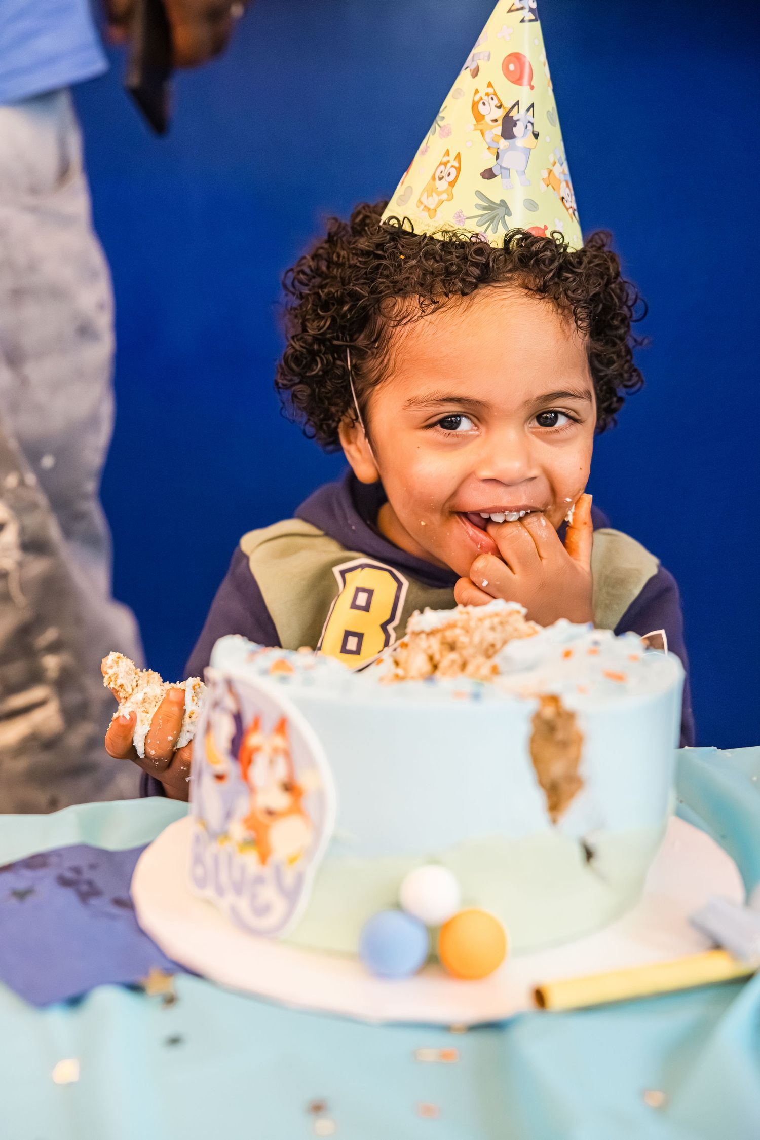 Happy young boy wearing party hat smiles while eating birthday cake with Bluey theme decoration.
