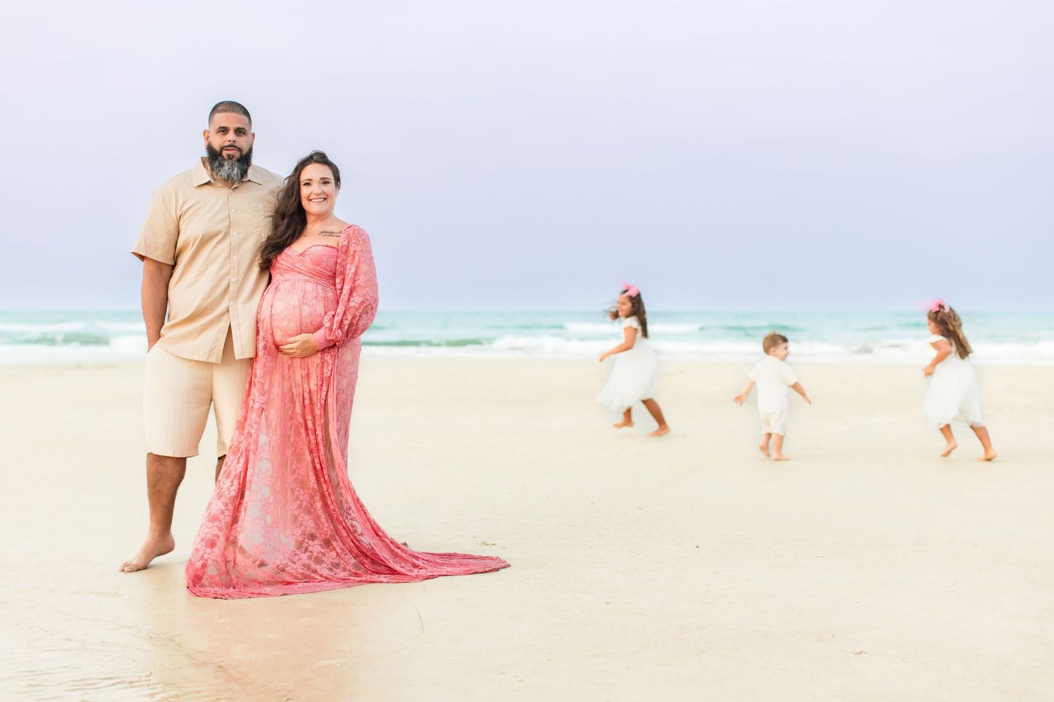 Pregnant woman in pink gown with partner on beach, children playing in background near ocean waves.