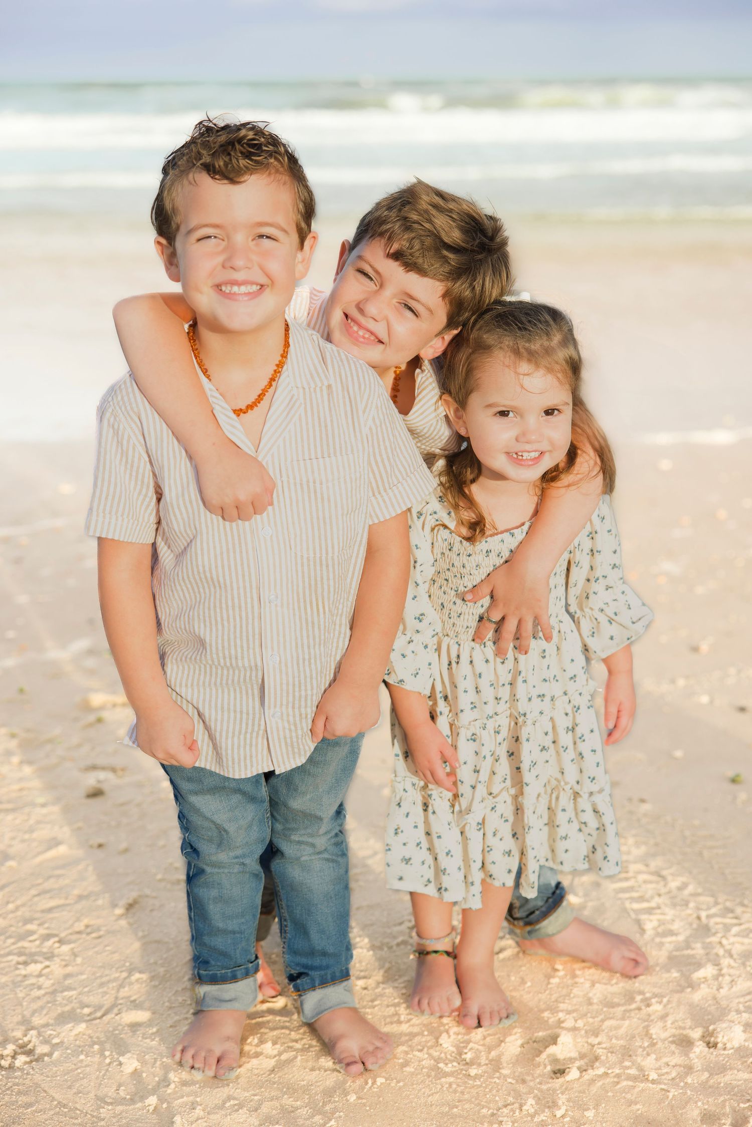 Three happy children hugging on a sunny beach, two boys and a girl smiling at the camera.