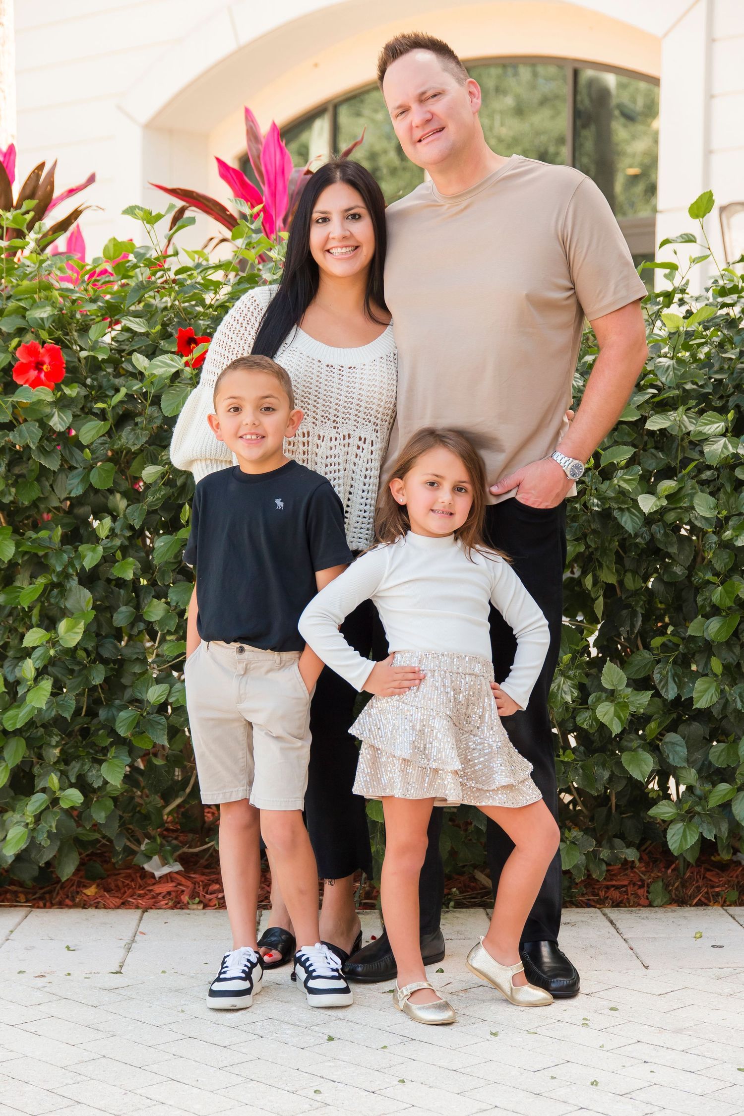 Family of four smiling together outdoors near colorful tropical flowers, parents with young boy and girl.