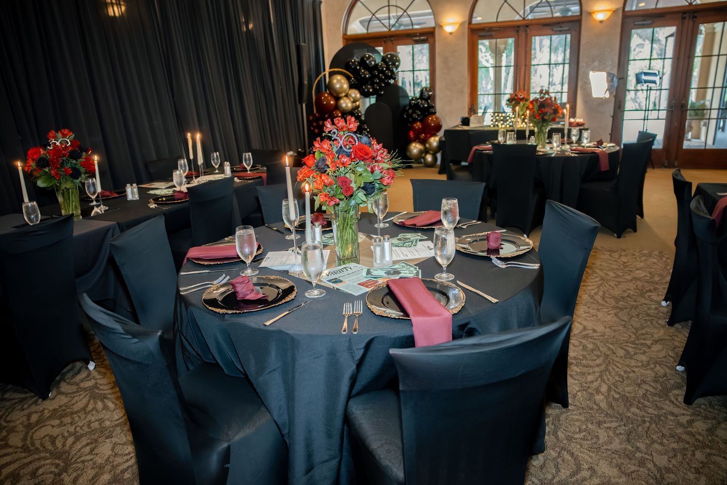 Formal event venue with navy blue tablecloths chair covers and vibrant red floral centerpieces on round tables.