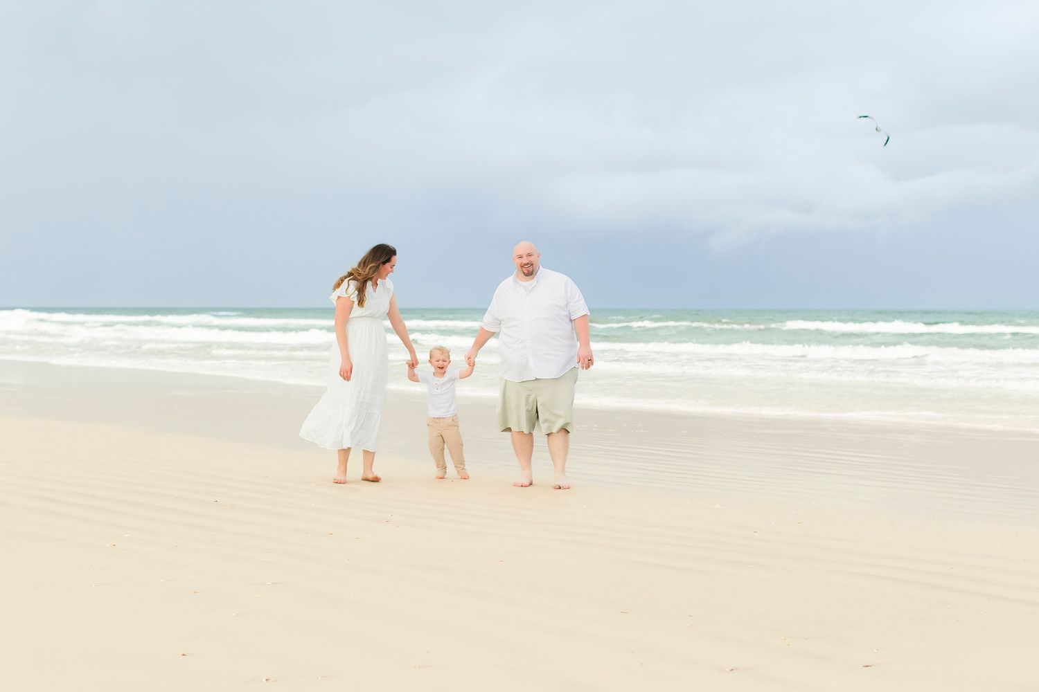 Young family of three playing on a white sand beach with turquoise ocean waves in the background.