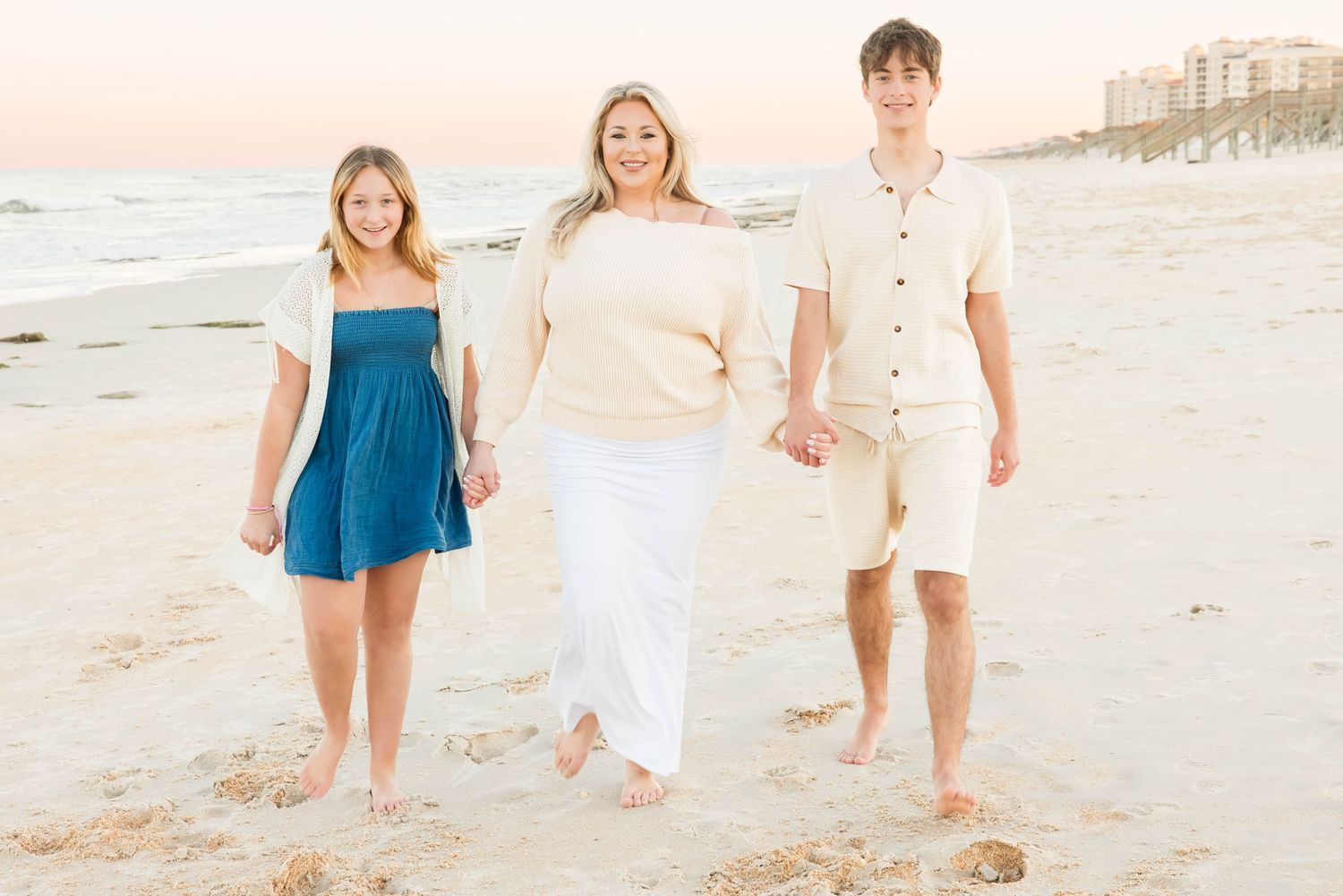 Mother holding hands with teenage son and daughter walking barefoot on sandy beach at sunset.