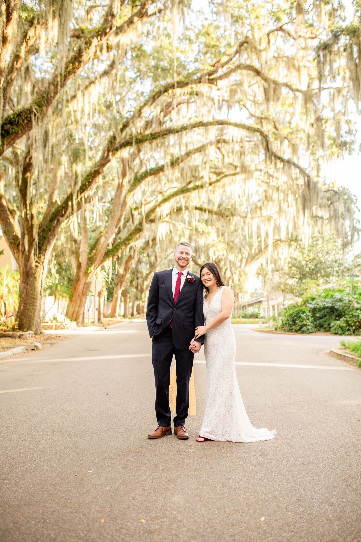 Smiling wedding couple holding hands on road beneath canopy of Spanish moss-draped oak trees.