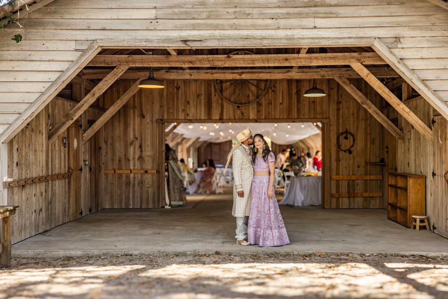 Couple in traditional Indian wedding attire stands in rustic barn wedding venue entrance.
