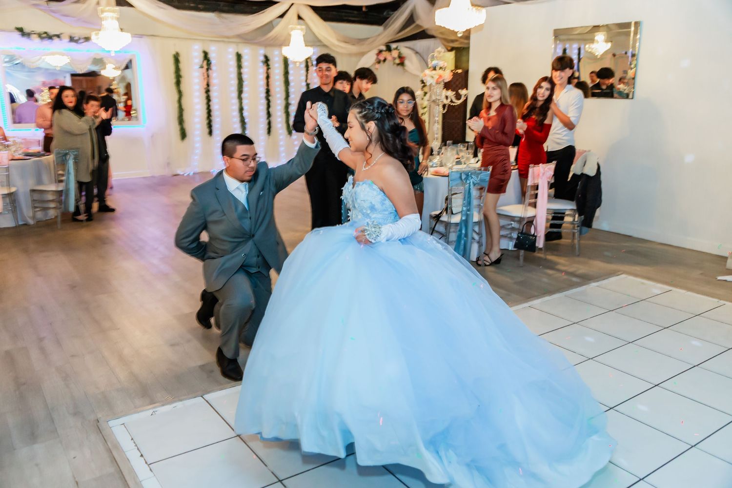 A girl in a blue ball gown dances with a partner at a lively quinceañera party.