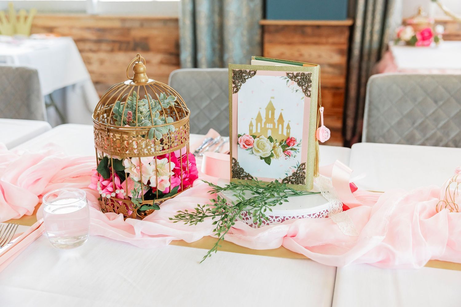 Elegant birthday party centerpiece with gold birdcage pink florals and decorative book on white tablecloth.