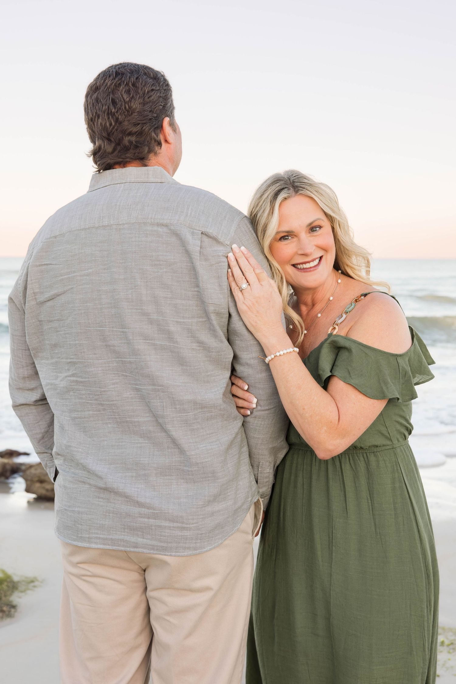 Smiling blonde woman in green off-shoulder dress embracing man from behind on beach.