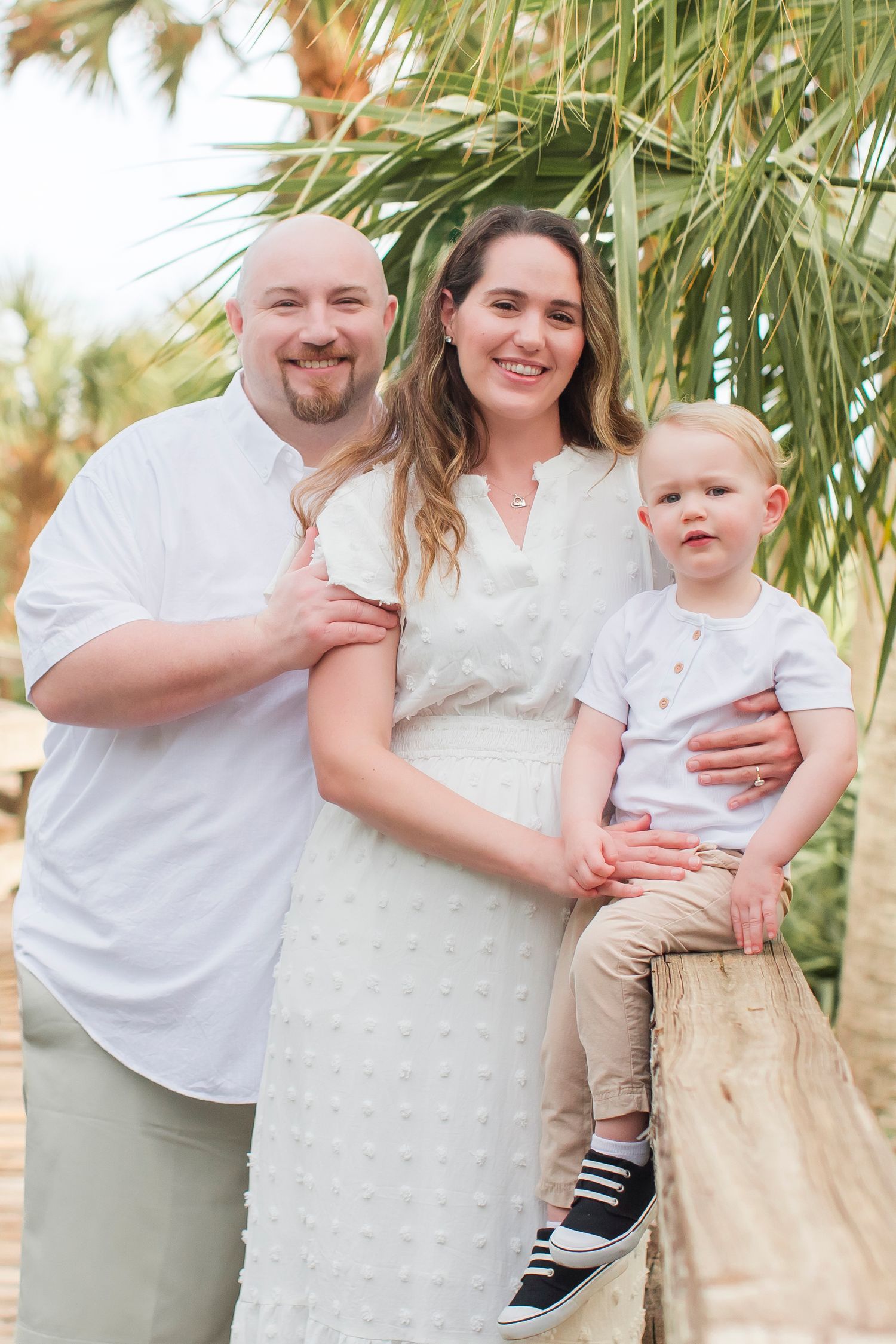 Smiling family portrait with bald father, brunette mother in white dress, and young toddler son outdoors.