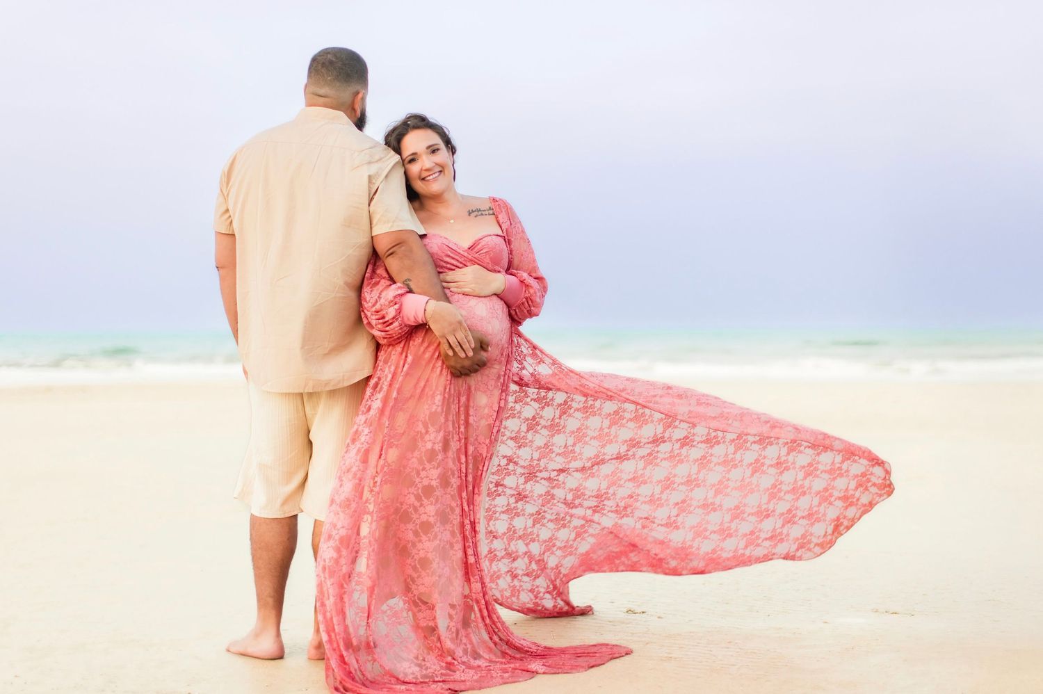 Pregnant woman in flowing pink dress poses with partner on a beautiful sandy beach.