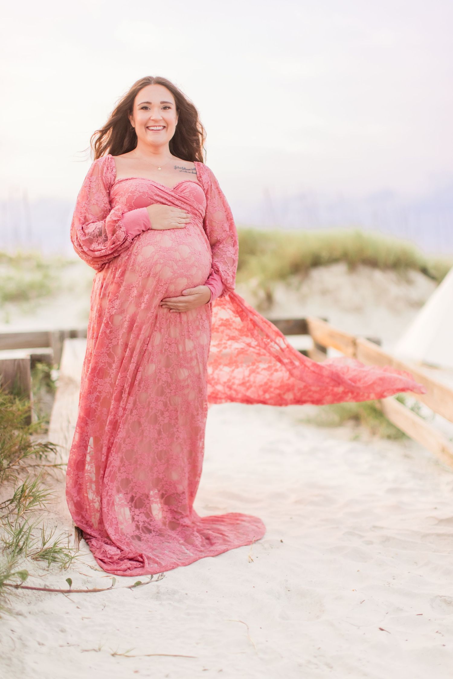 Pregnant woman in flowing pink lace maternity gown posing on sandy beach dunes at sunset.
